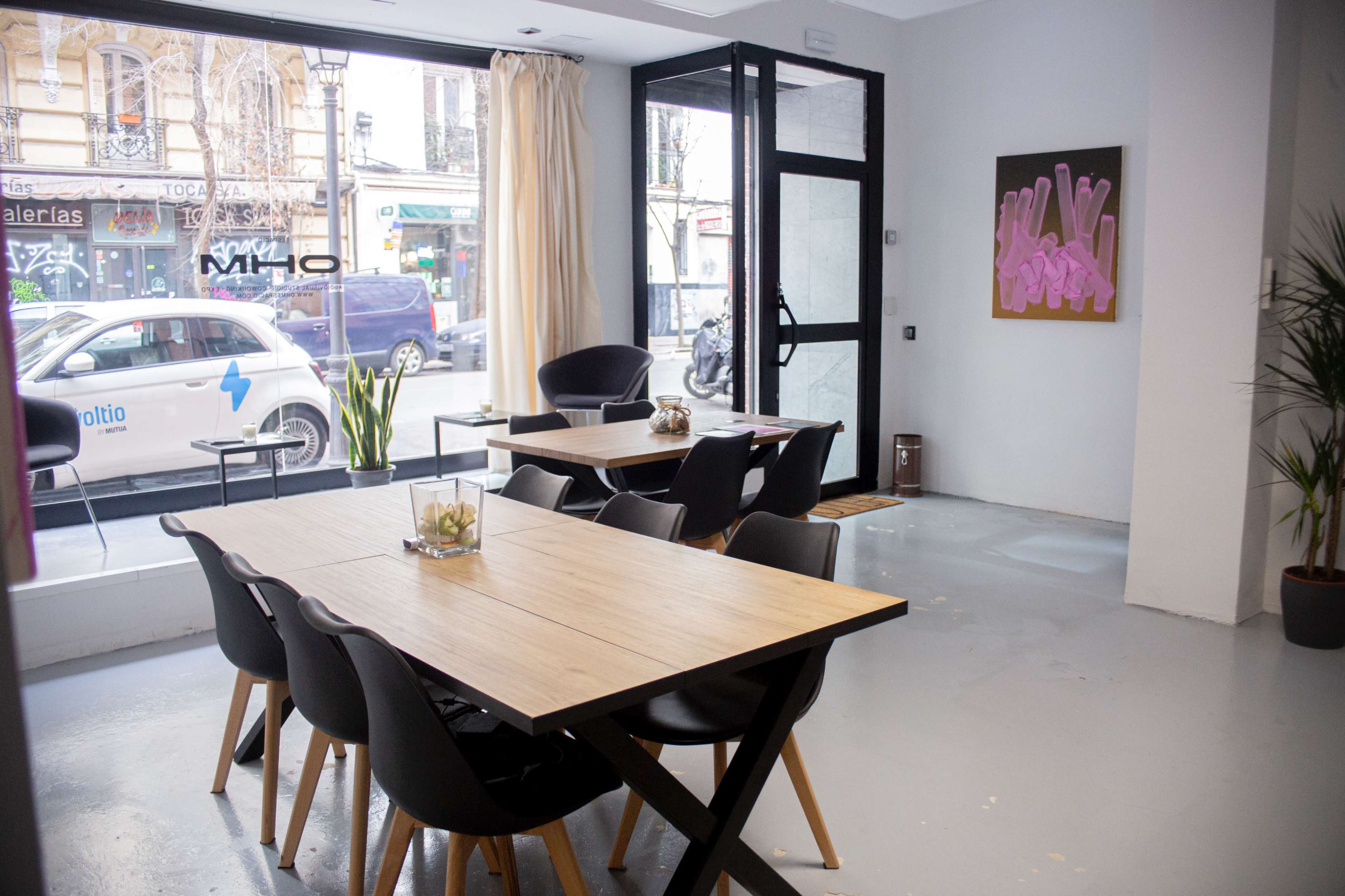 The image shows a modern interior of a café or restaurant with wooden tables, black chairs, and a large window facing a street.