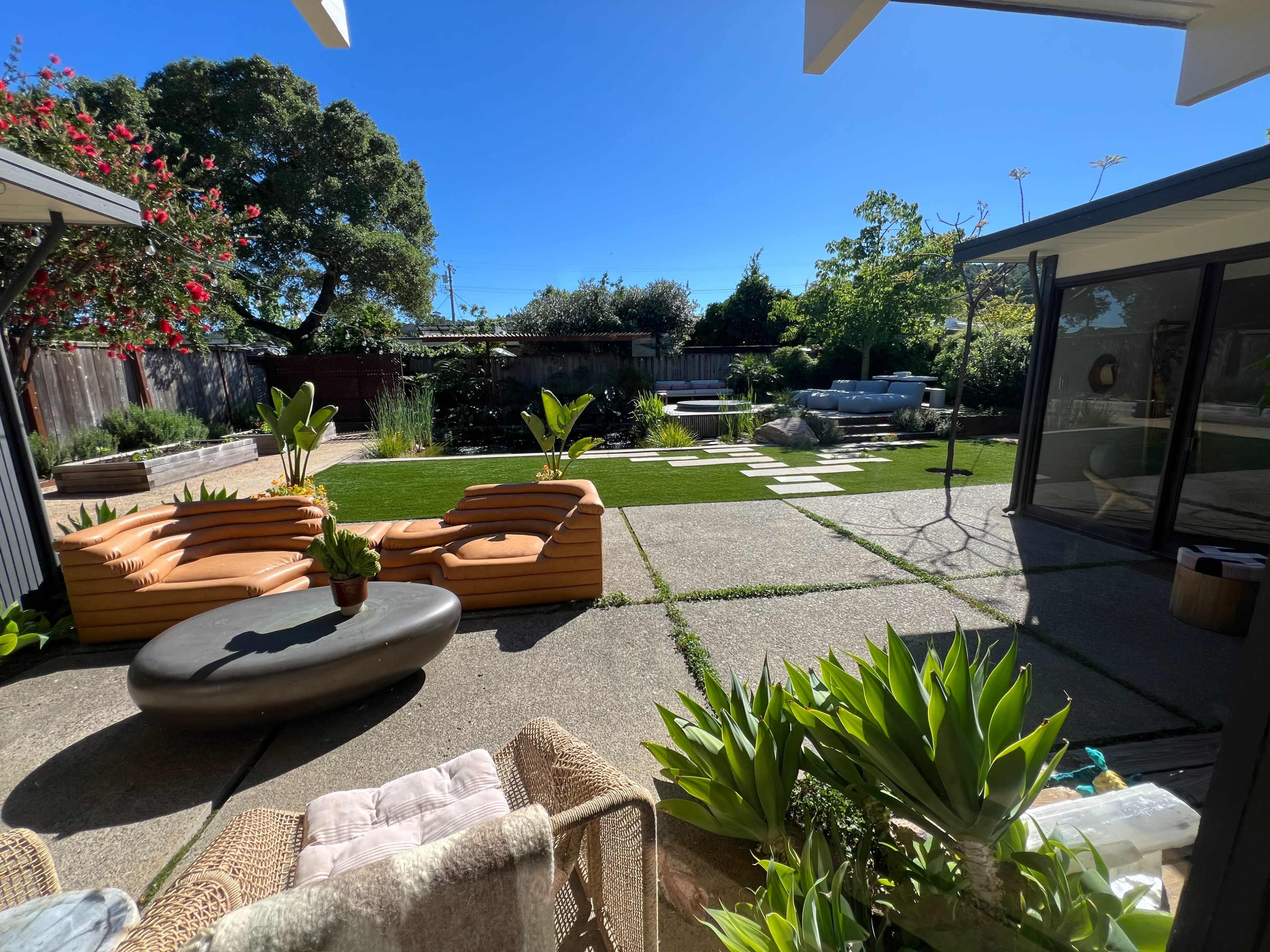 The image shows a landscaped backyard with a seating area made of wooden couches, a round coffee table, and a garden featuring greenery and a stone path.