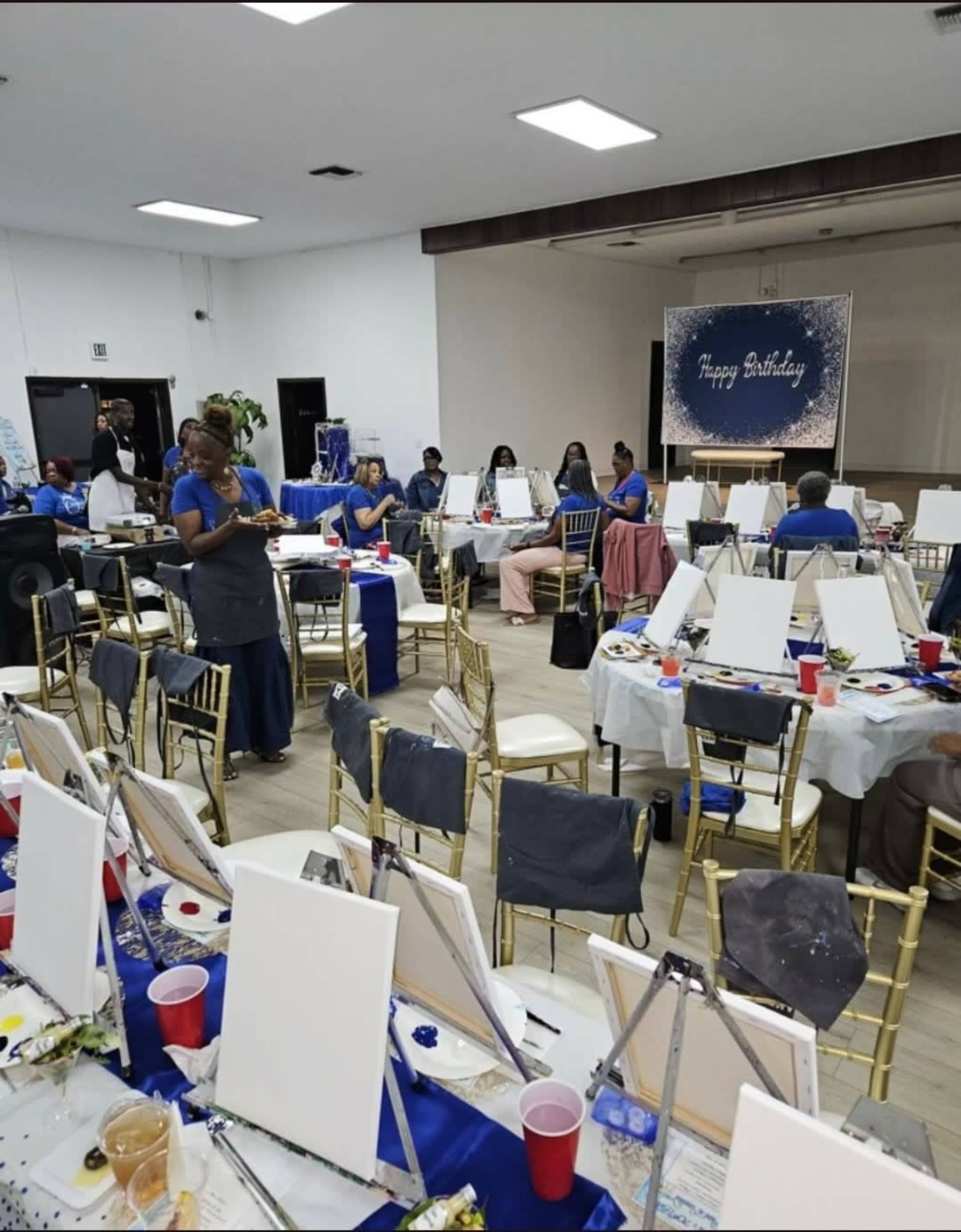 The image shows a birthday celebration in a spacious room, with tables set up for painting activities, each adorned with blank canvases, paint supplies, and blue decorations.