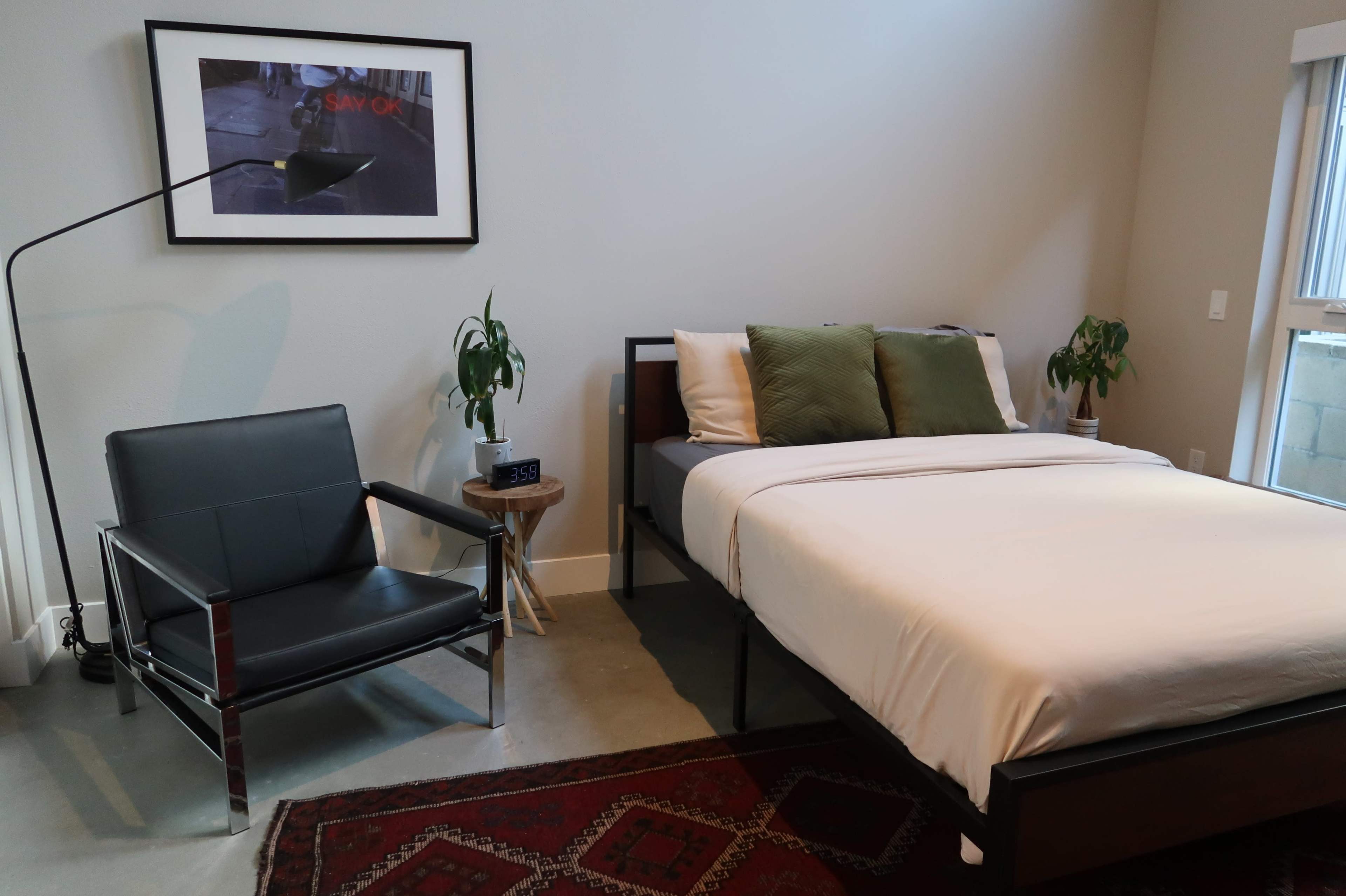 The image shows a minimalist bedroom featuring a bed with a light-colored bedspread, a black chair next to a small table, and a framed picture on the wall.