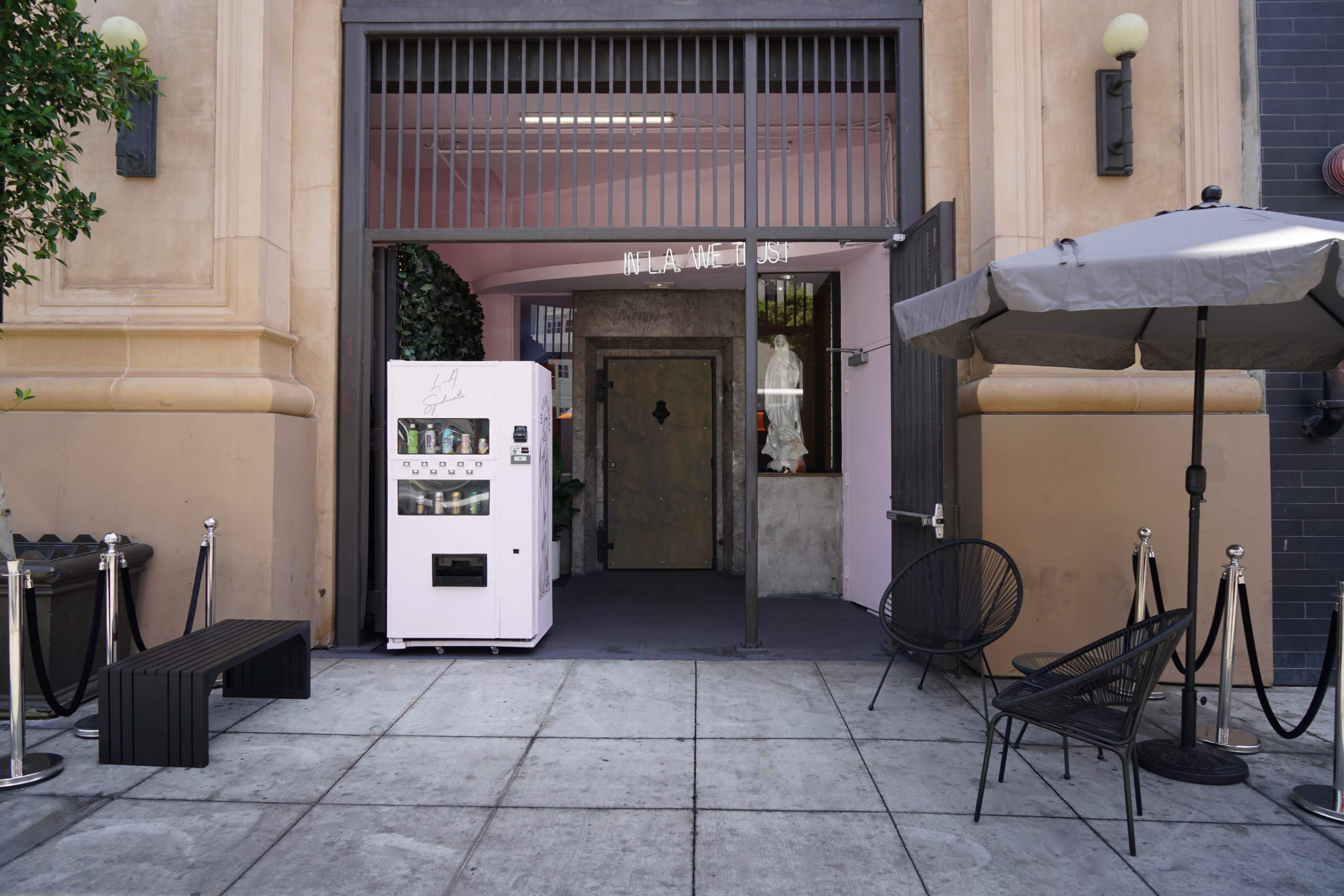 The scene features a simple entrance with a pink wall, a white vending machine near a large door, and a few outdoor seating arrangements.