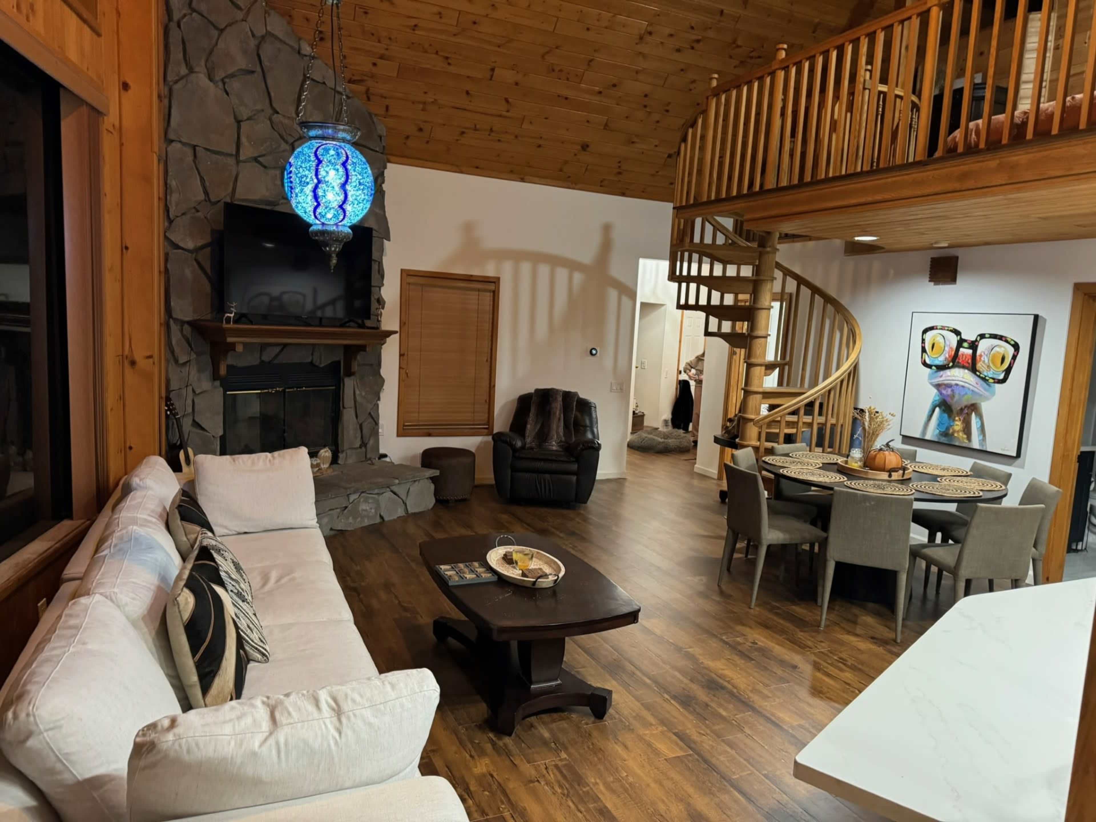 The image shows a spacious, rustic interior of a cabin with a stone fireplace, a spiral staircase, and a dining area with a large round table.