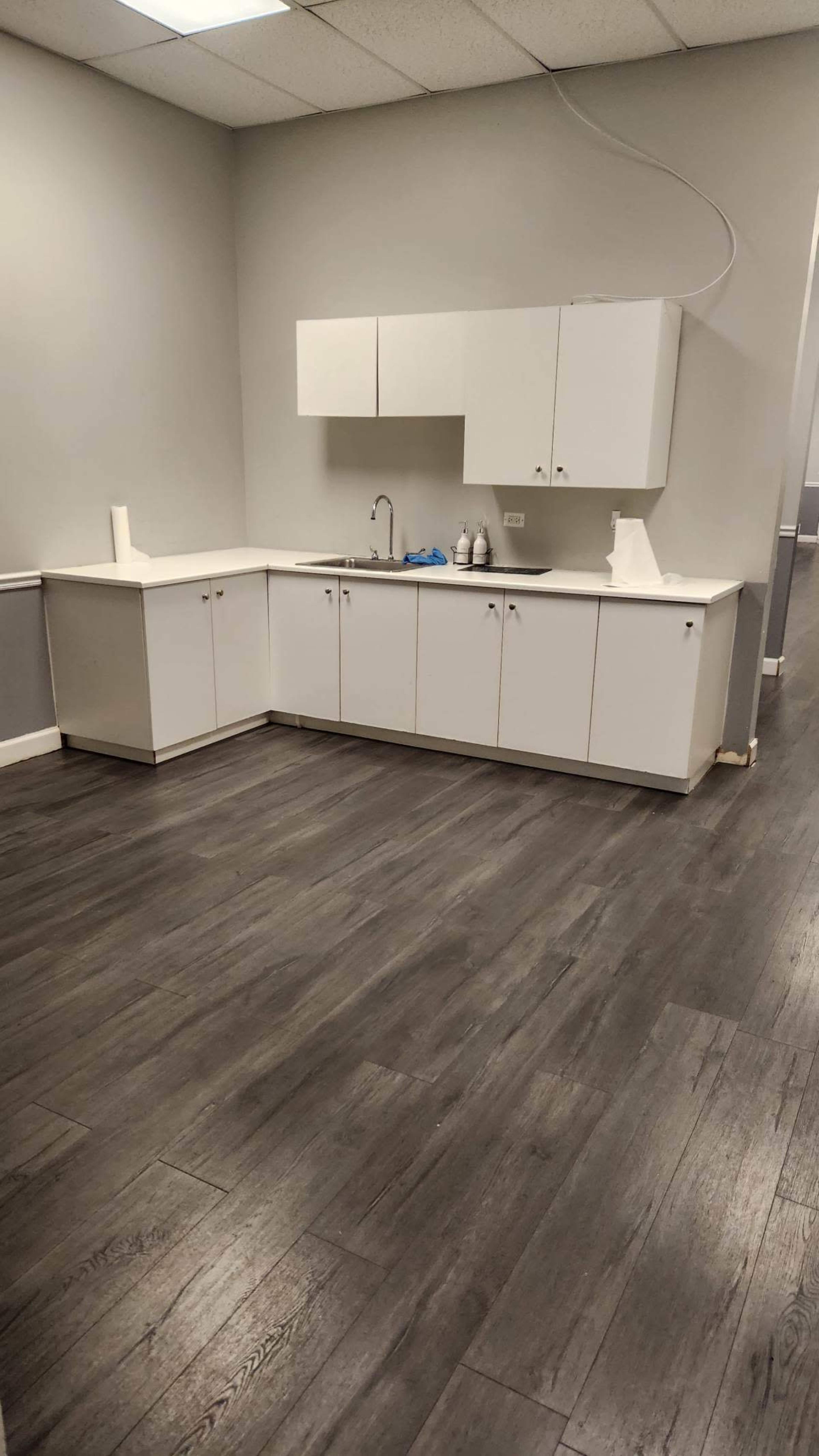 The image shows a clean, empty kitchen area with white cabinets, a sink, and gray laminate flooring.