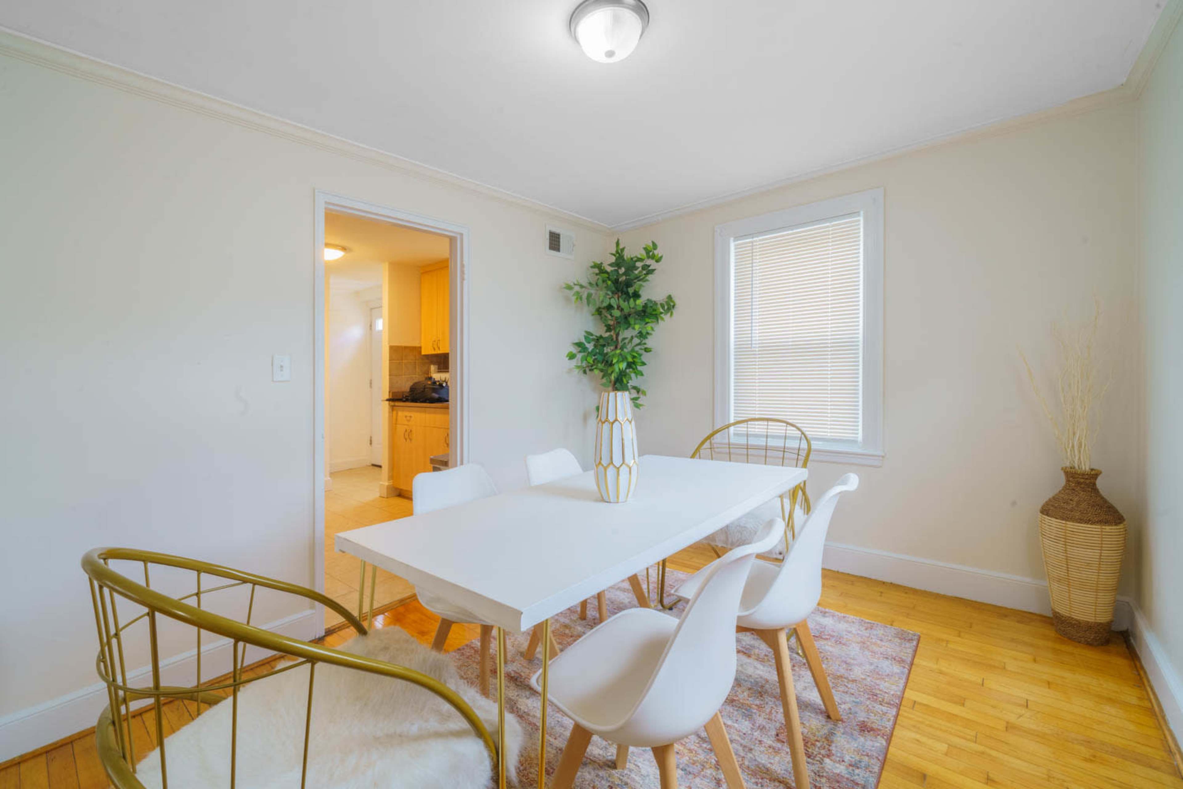 A dining area features a white table surrounded by four chairs, with a potted plant as a centerpiece and a doorway leading to a kitchen.