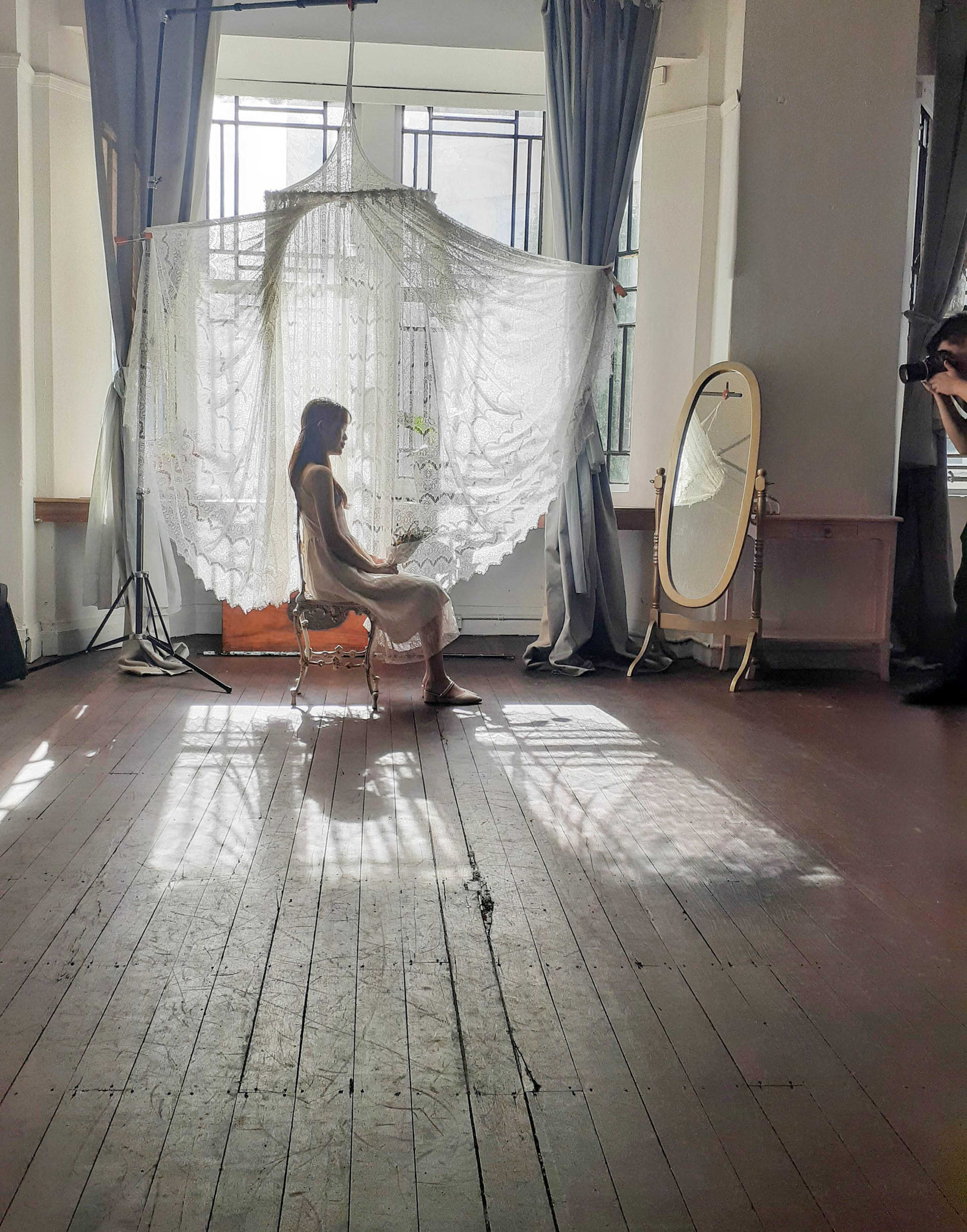 A woman sits on a chair in a well-lit room with a large lace curtain and an ornate mirror nearby, casting intricate shadows on the wooden floor.