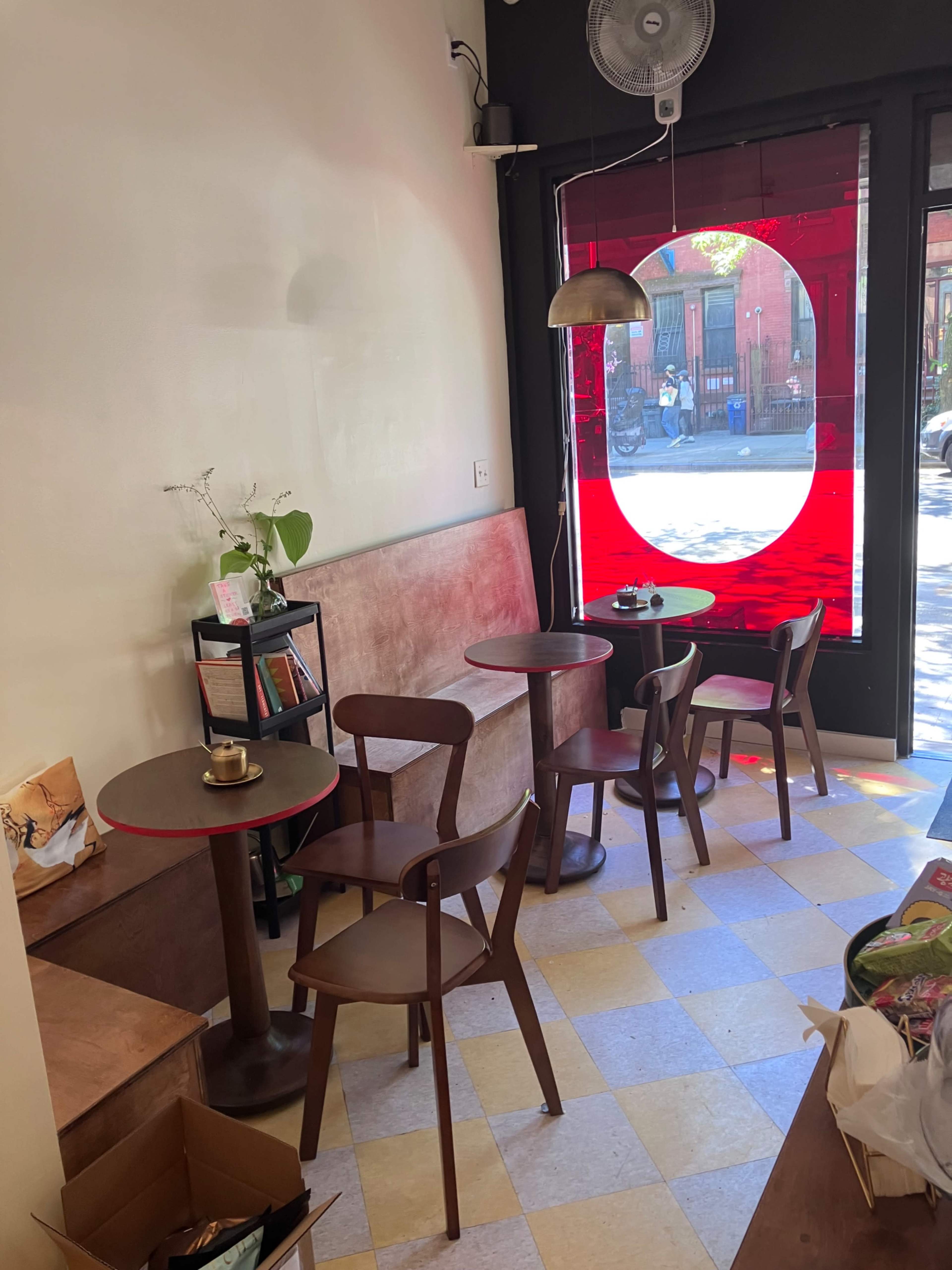 The image shows a small café interior with a few wooden tables and chairs, a large red circular window, and a fan mounted on the wall.