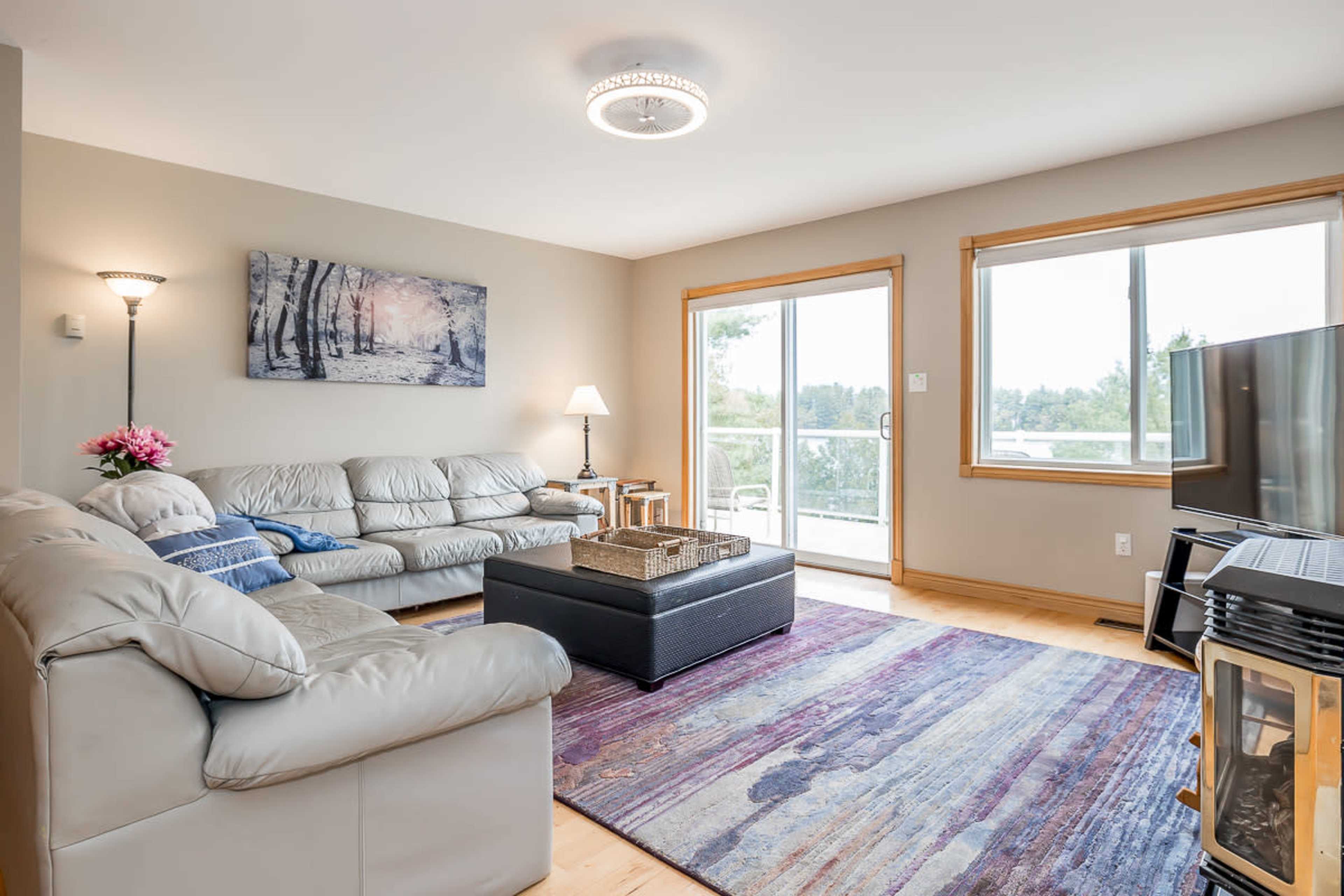 The living room features a light-colored sofa set, a coffee table on a patterned rug, and large windows providing natural light.