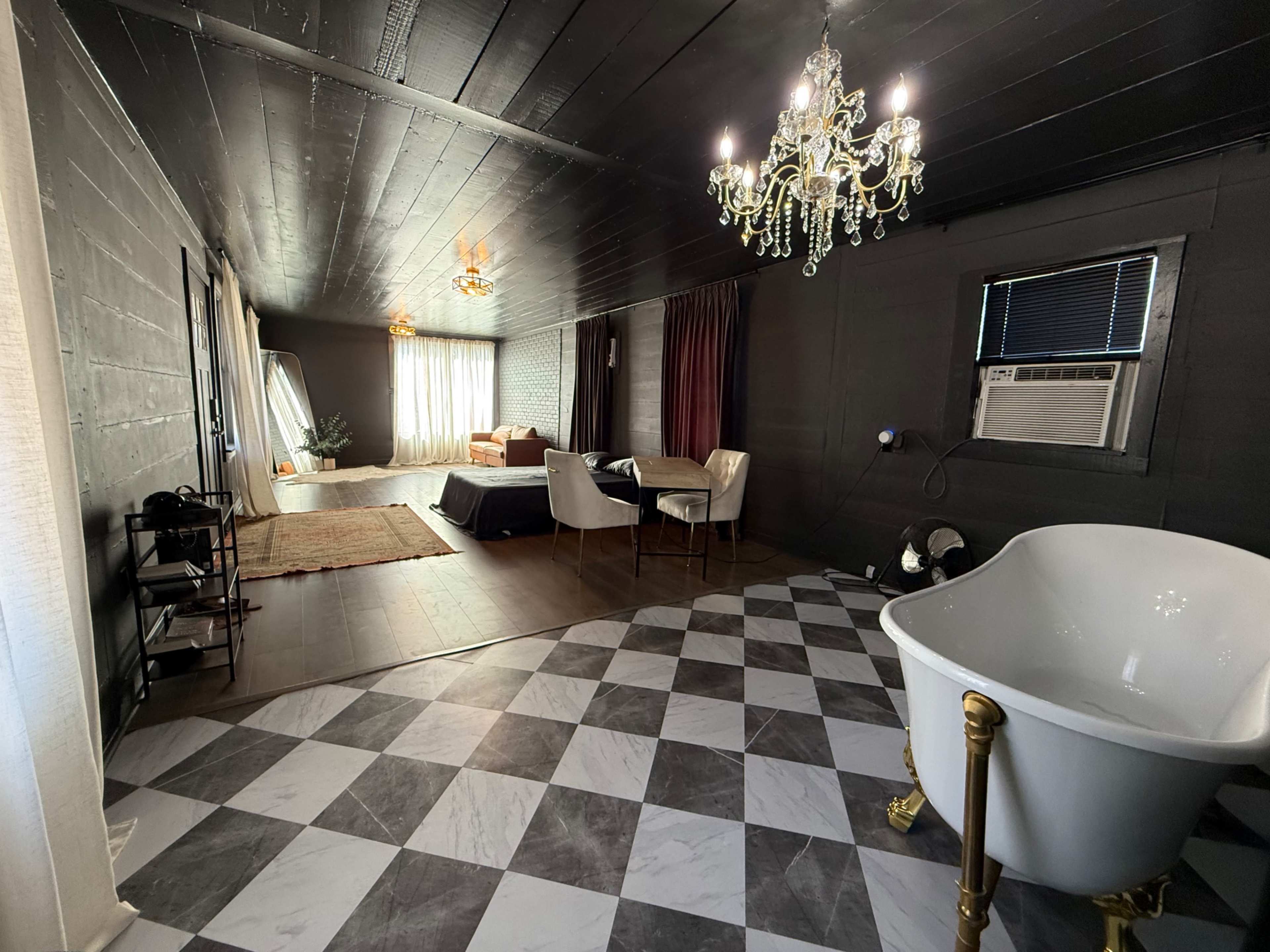 The image shows a spacious room with dark walls, a large window allowing natural light, a chandelier, a freestanding bathtub, and a checkered floor pattern.