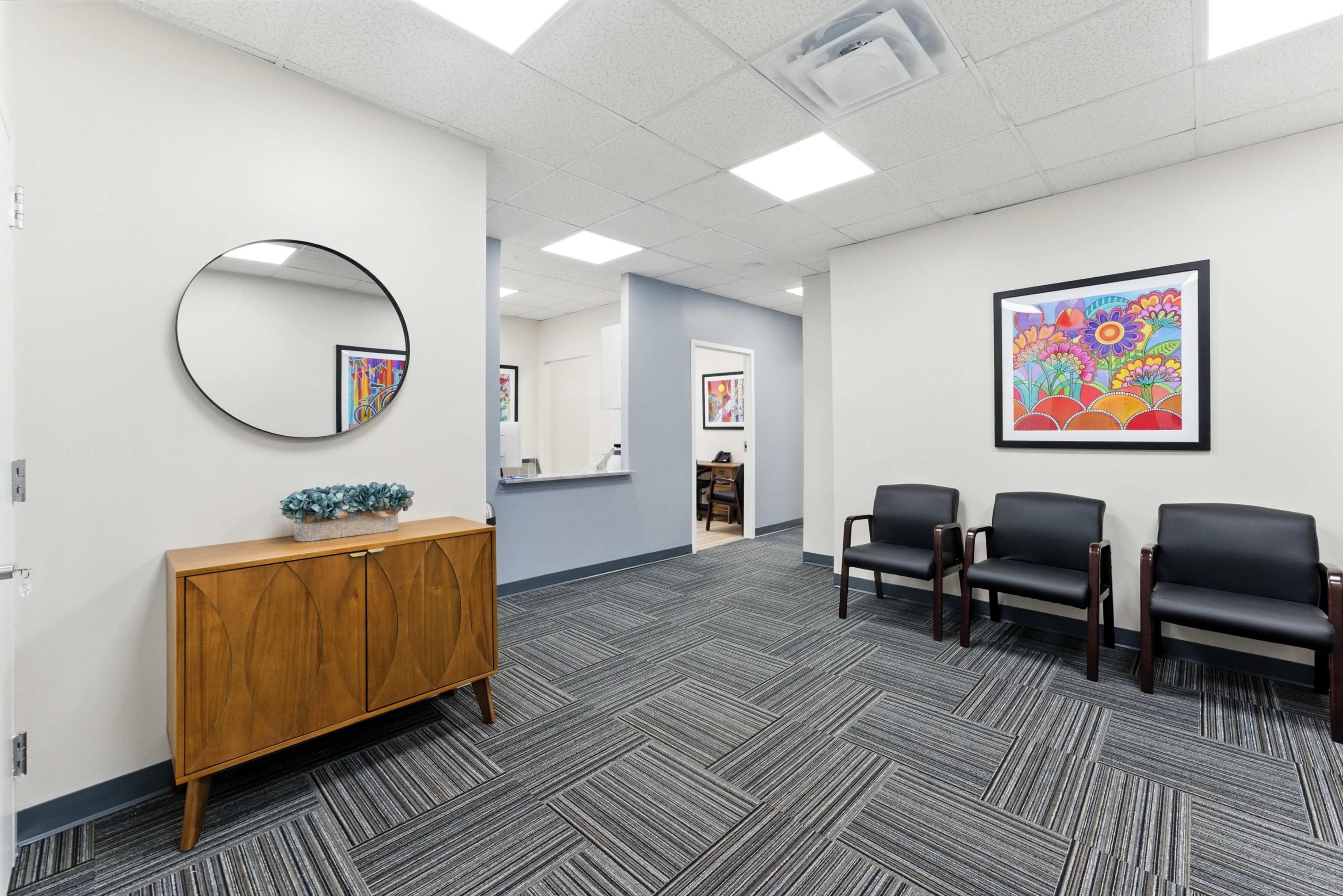 The image shows a waiting area with a wooden cabinet, black chairs, and colorful framed artwork on the walls.