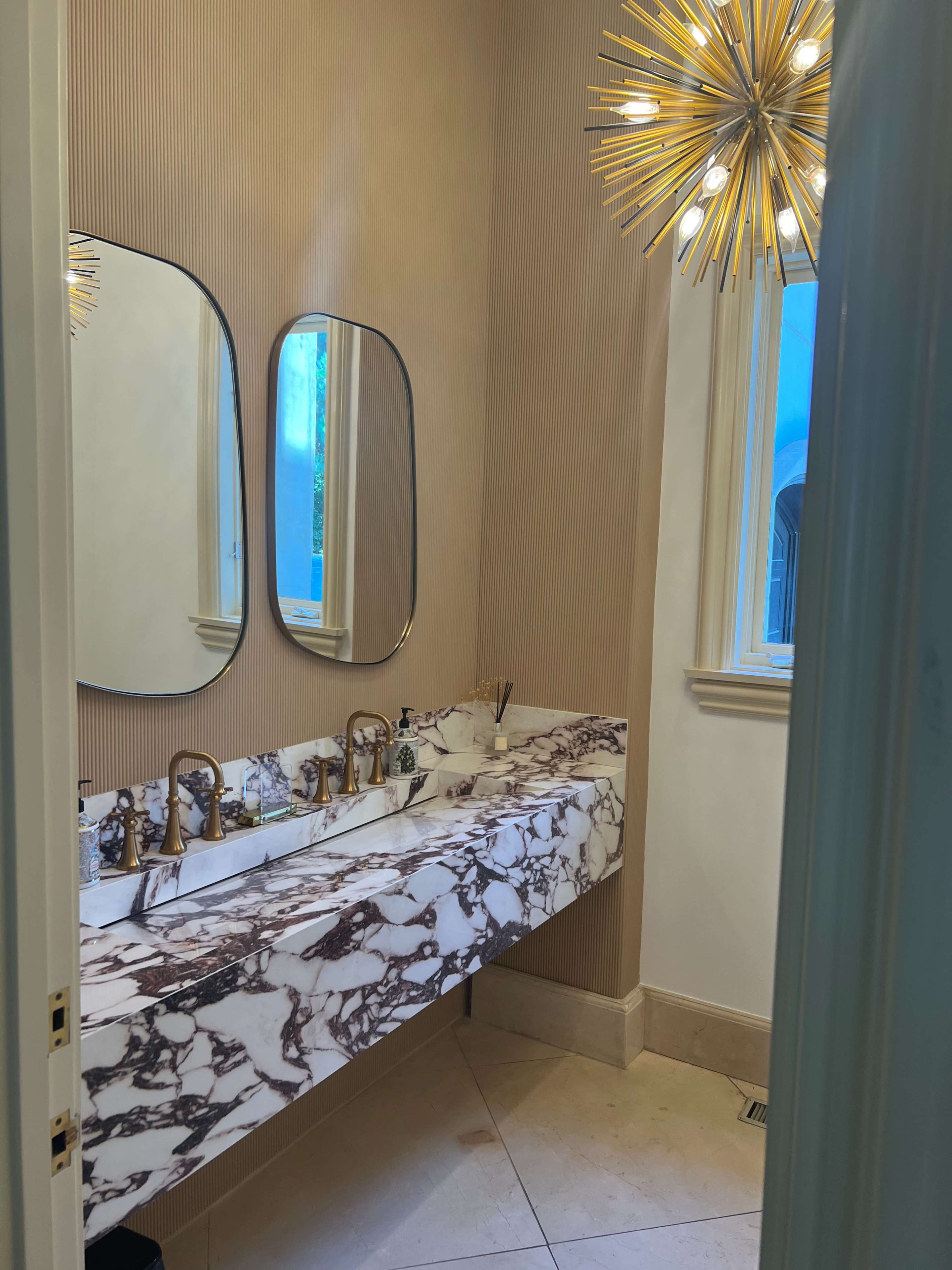 The image shows a modern bathroom with a marble vanity featuring two mirrors and multiple faucets, illuminated by a starburst chandelier.