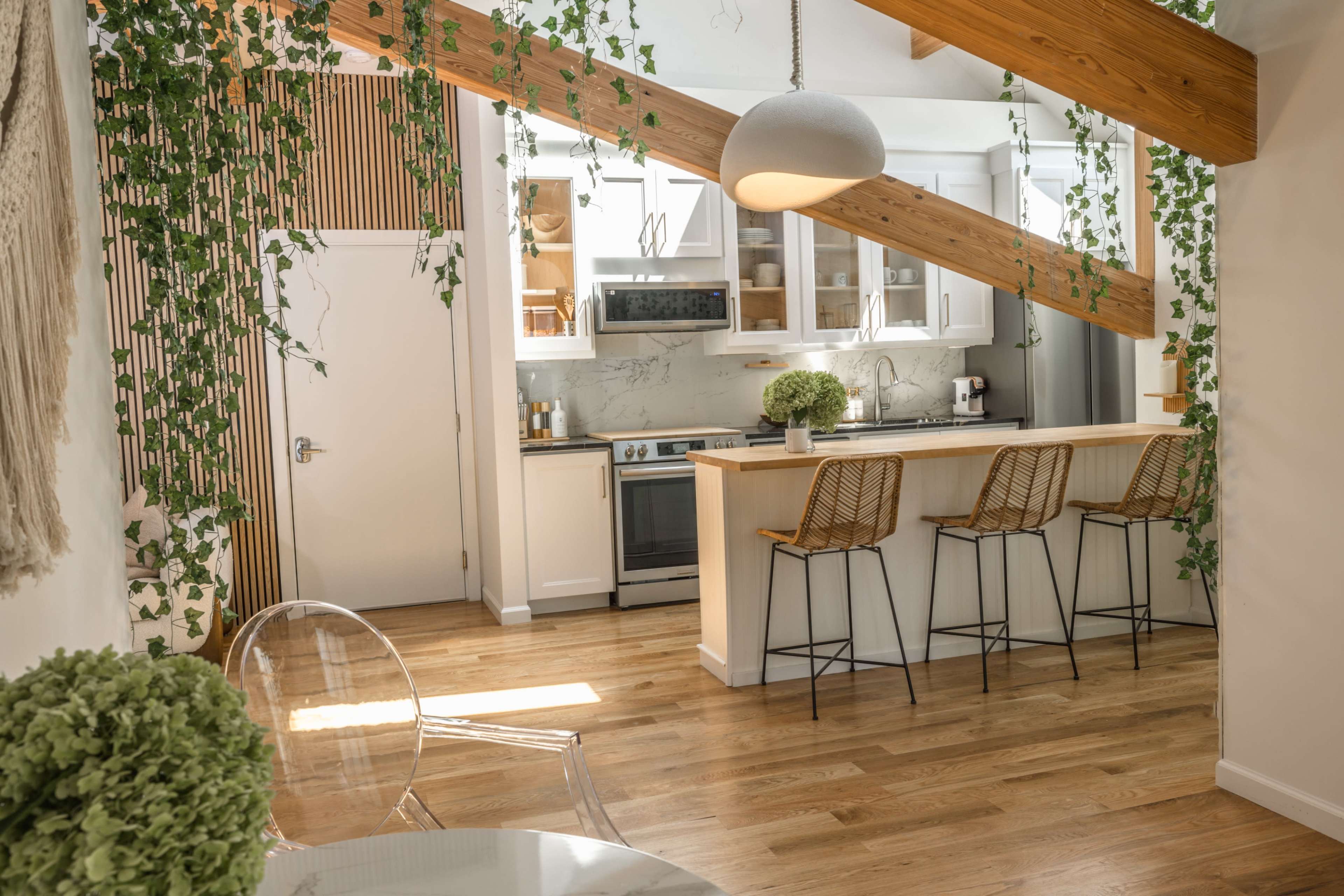 A modern kitchen with wooden beams, white cabinetry, and bar stools, accented by greenery and natural light.