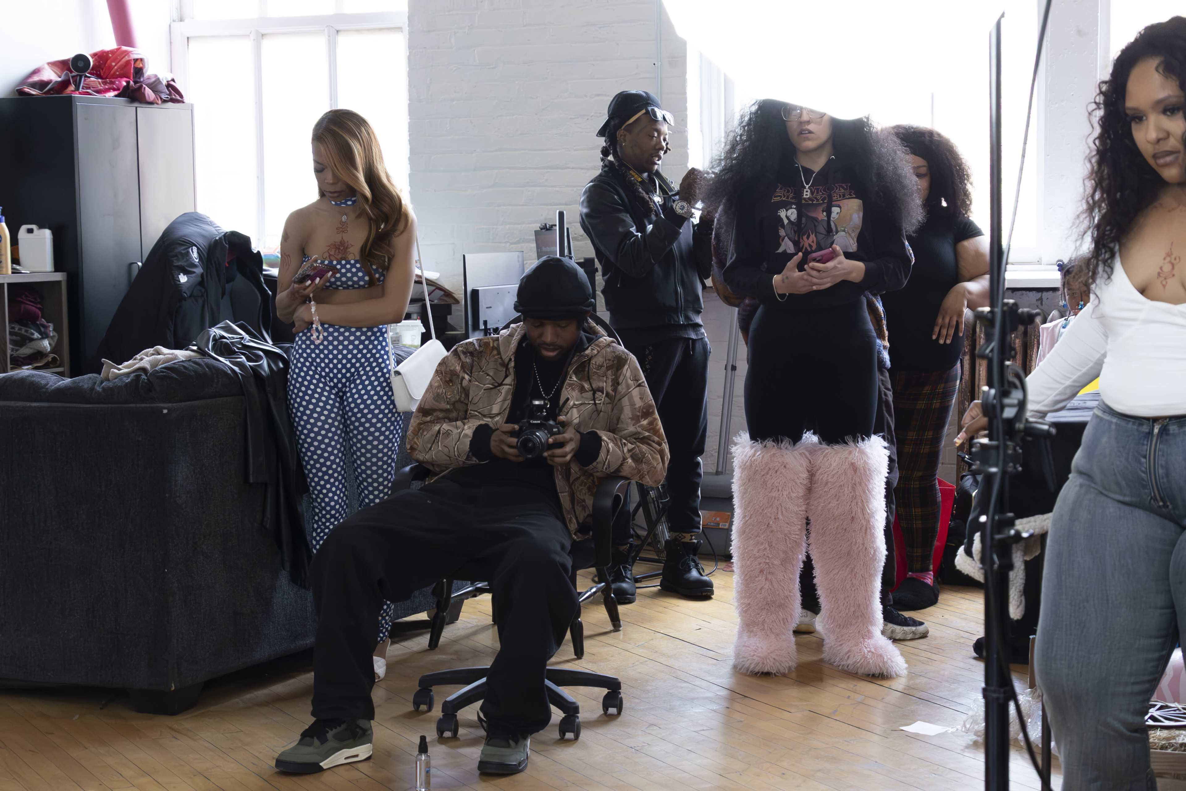A group of individuals is gathered in a bright studio, with some standing and others seated, while a photographer operates a camera.