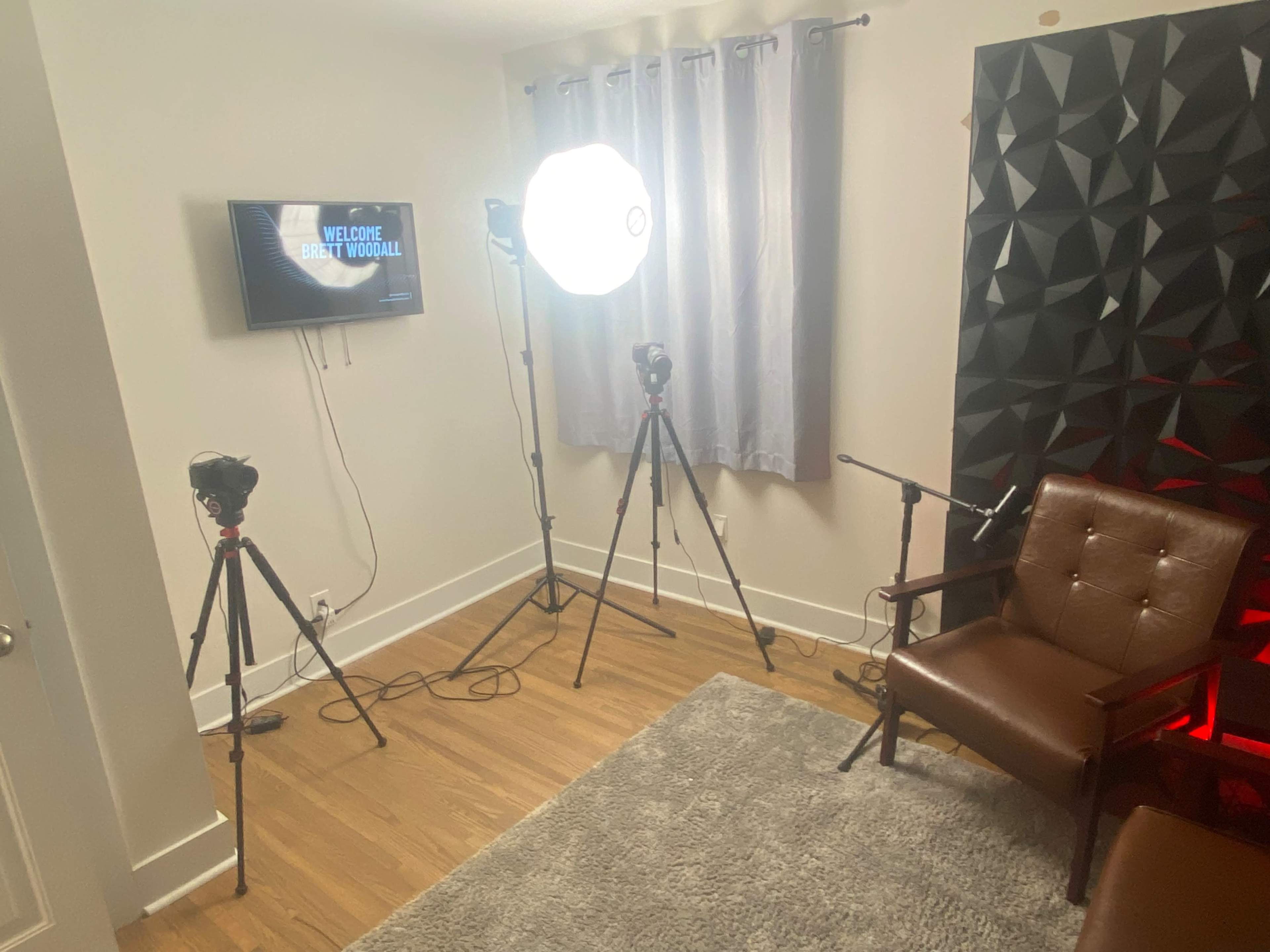 The image shows a small room set up for video recording, featuring two cameras on tripods, a softbox light, a brown armchair, and a wall display with a welcome message.