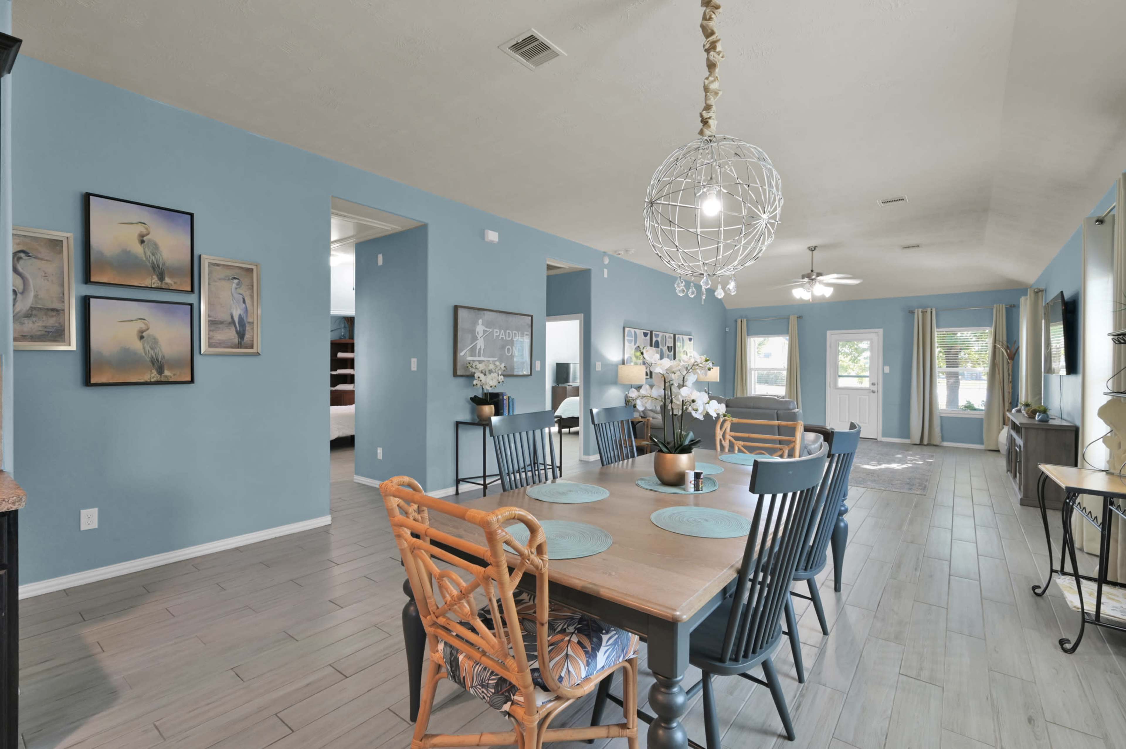 The image shows a spacious dining area with a large table surrounded by wooden chairs, blue walls, and decorative art on the walls.