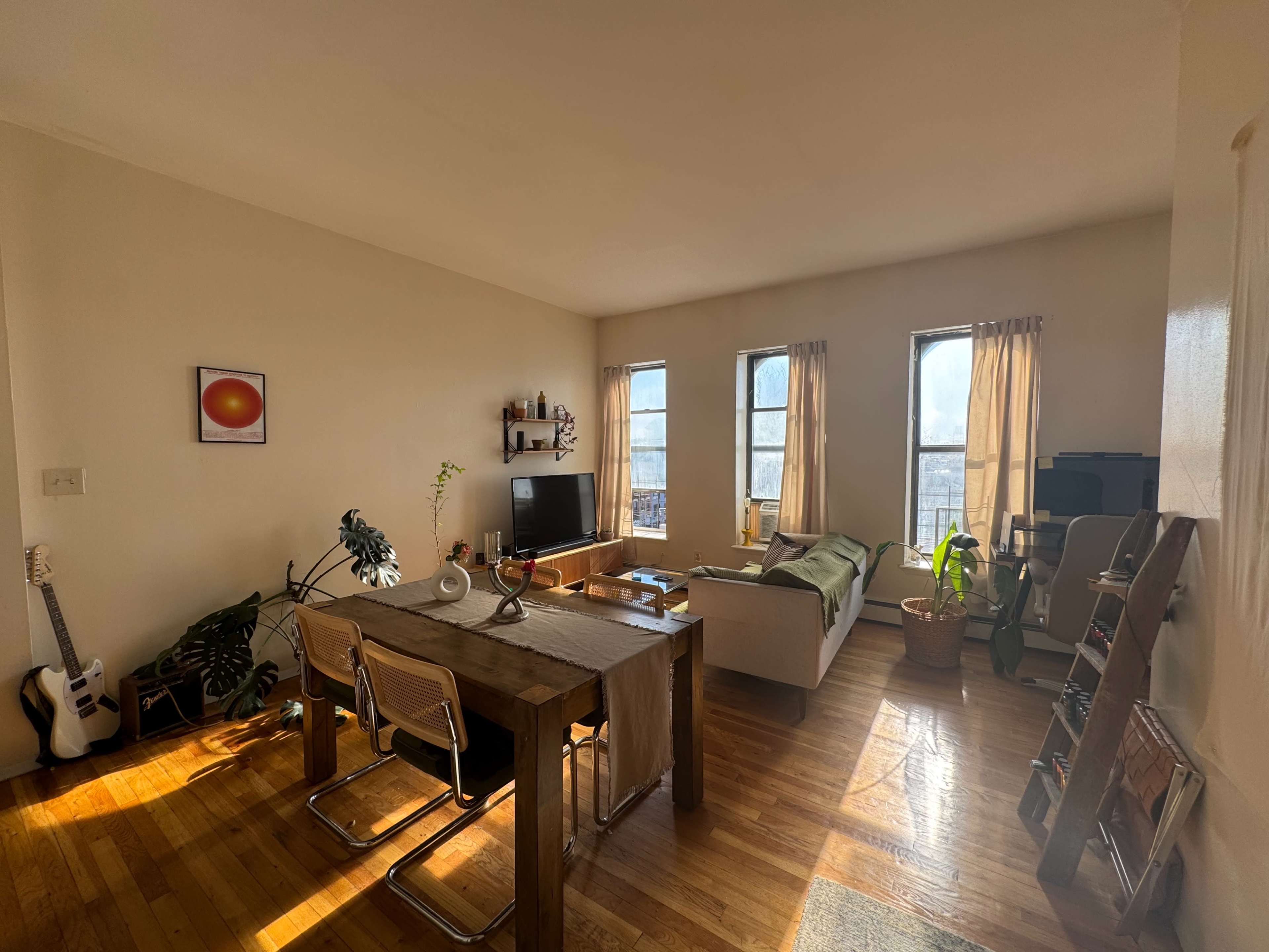 A bright living room features a sofa, dining table with chairs, and large windows allowing natural light to fill the space.