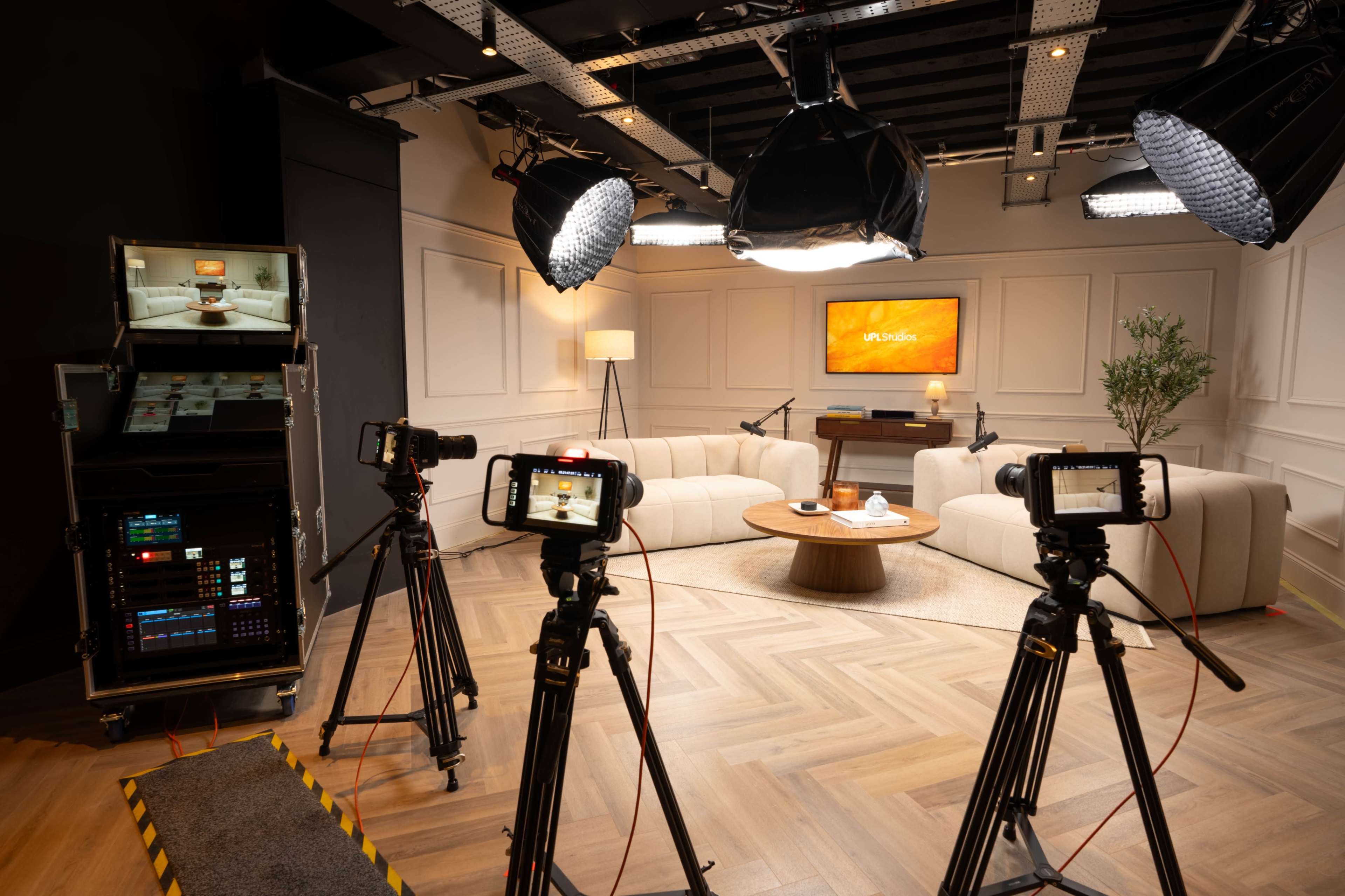 A modern television studio setup features three tripods with cameras facing a circular seating area, surrounded by bright lighting and a backdrop displaying the “UP Studios” logo.
