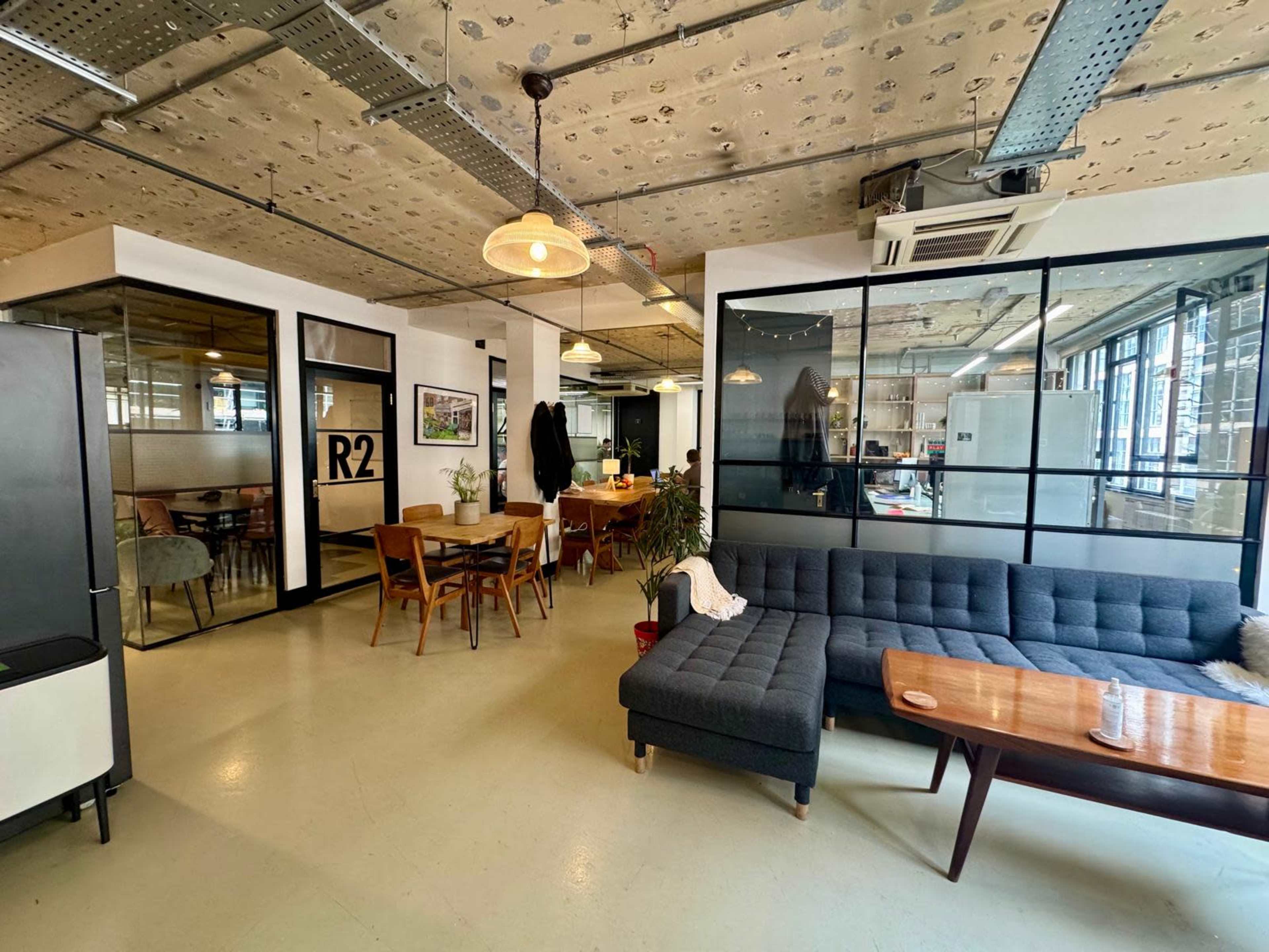 The image shows a modern open office space featuring a mix of lounge seating, dining tables, and glass-walled rooms.