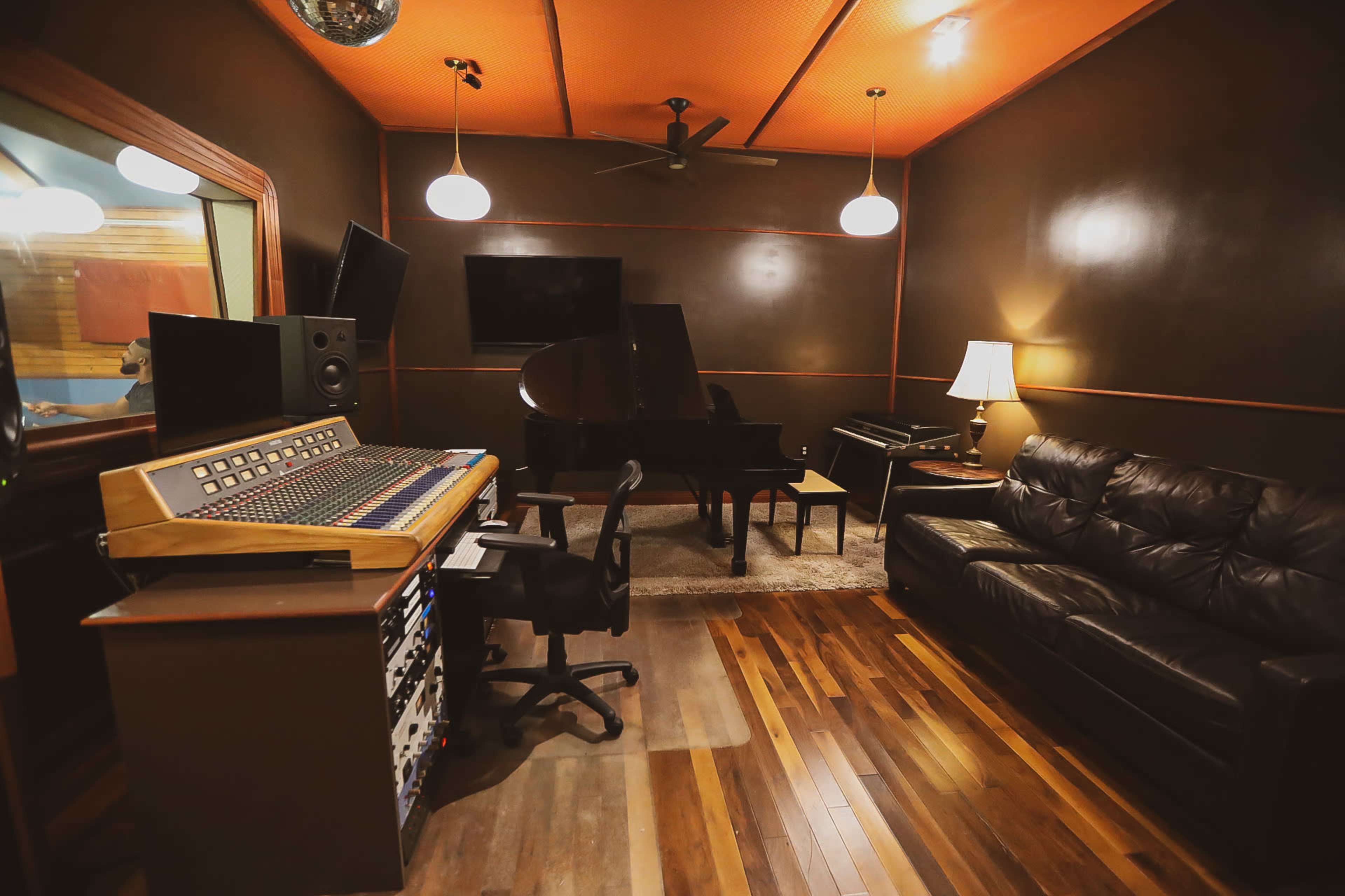 The image features a music studio with a mixing console, a grand piano, and a leather sofa in a dimly lit room.