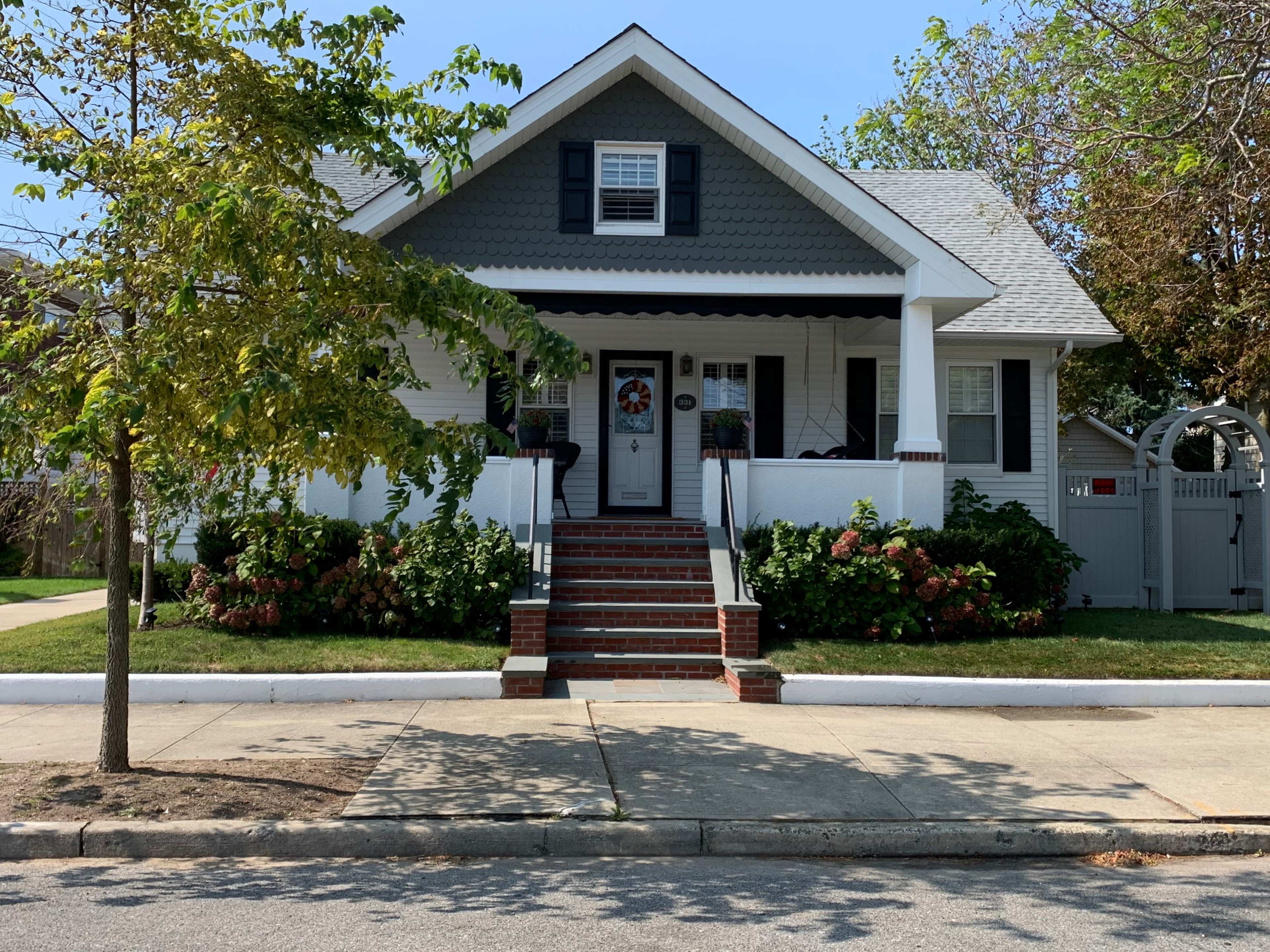 The image shows a two-story house with a gabled roof, white trim, and red brick steps leading to a front door surrounded by flower pots.