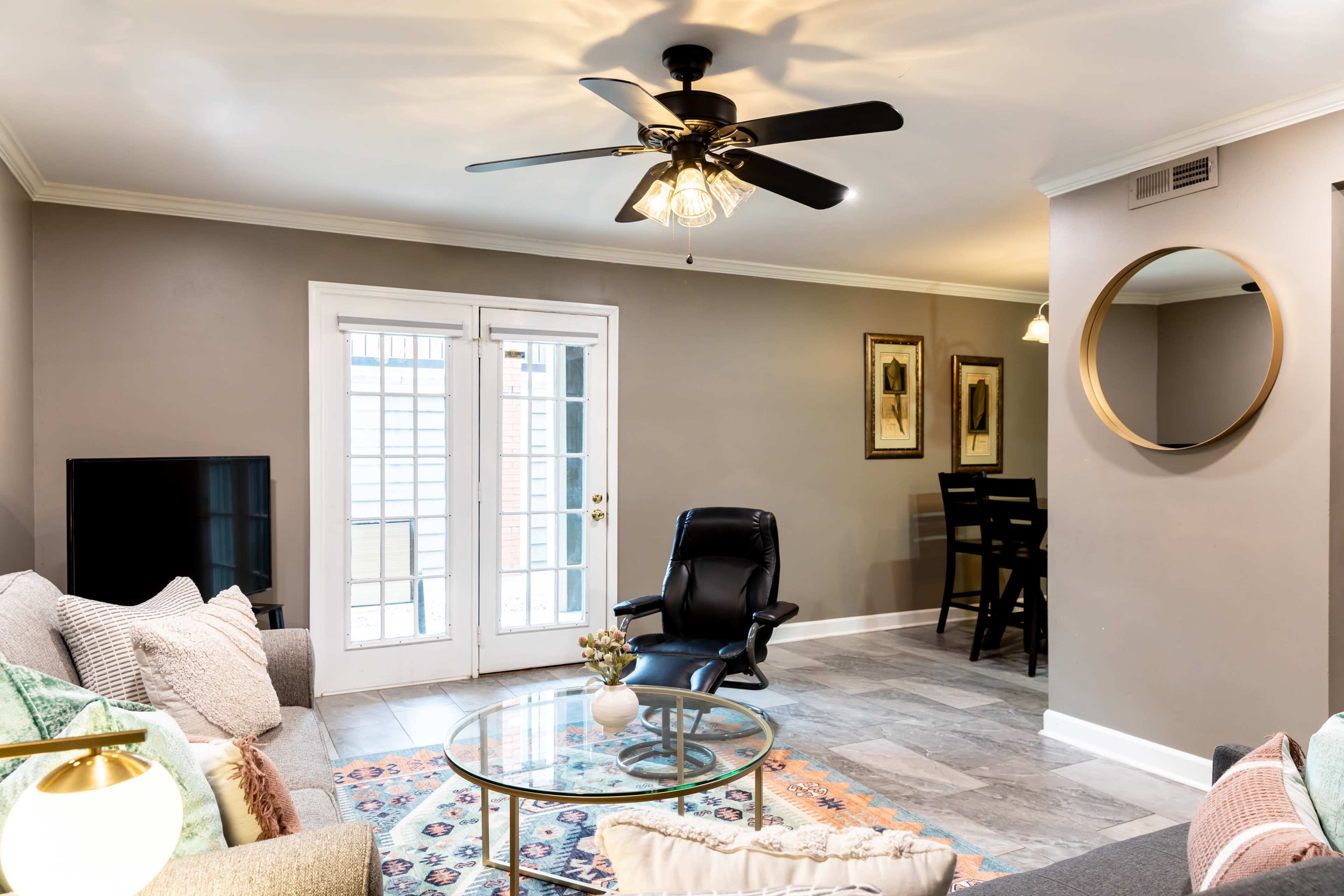 The image shows a modern living room with a ceiling fan, a black leather recliner, a glass coffee table, and a TV, with sliding glass doors leading to an outdoor area.