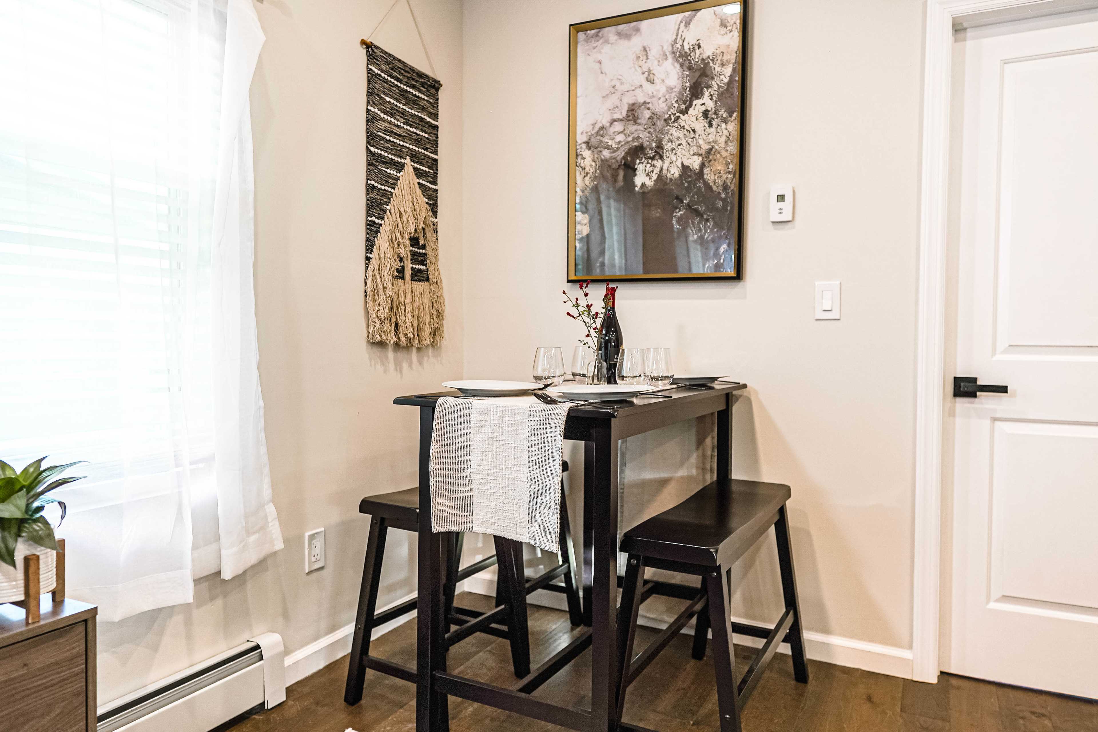 A small dining area features a black table set with glasses and a bottle of wine, accompanied by two dark stools, a wall hanging, and a framed artwork.