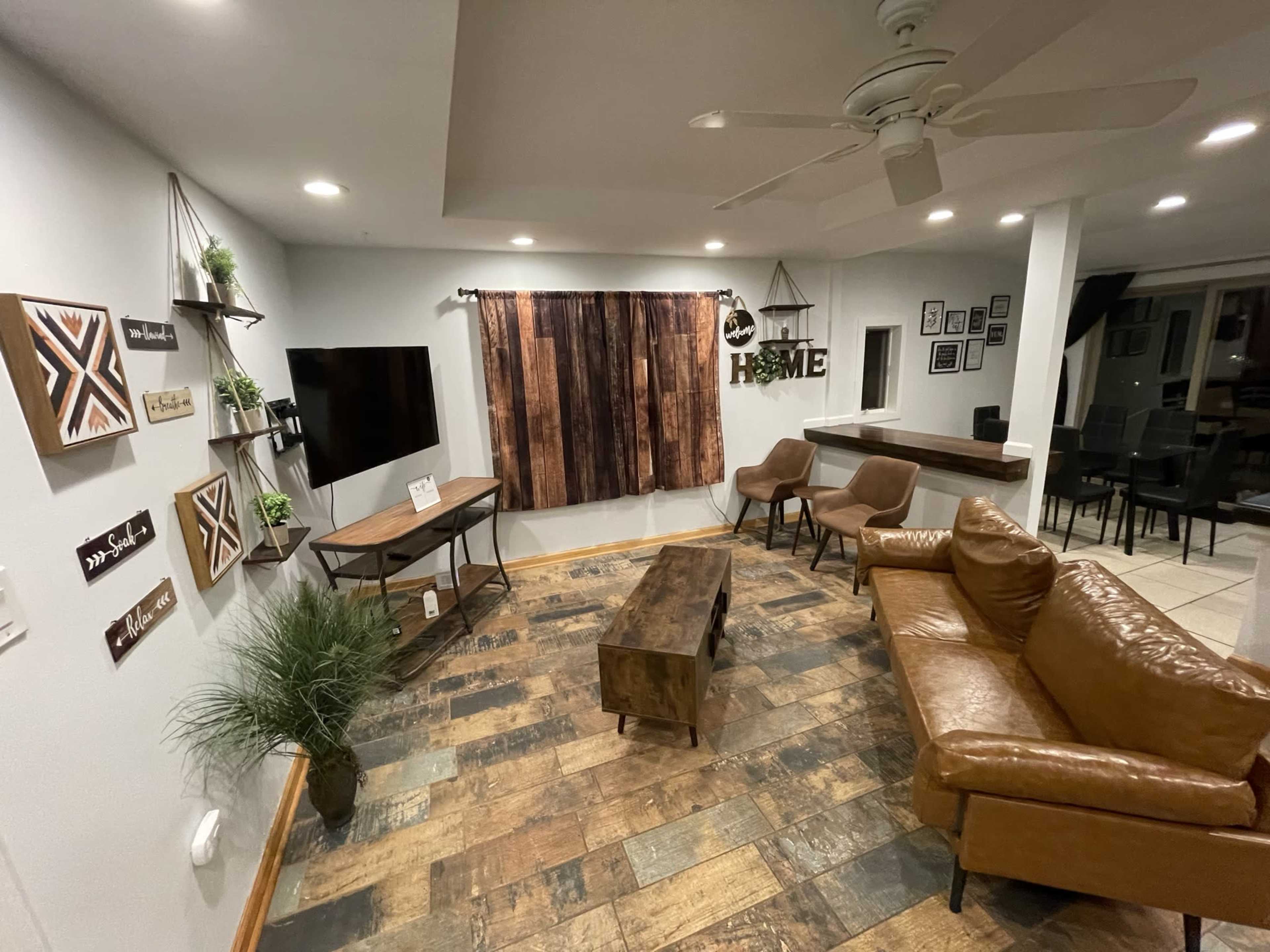 The living room features a leather sofa, two chairs, a wooden coffee table, a television mounted on the wall, and decorative wall art.