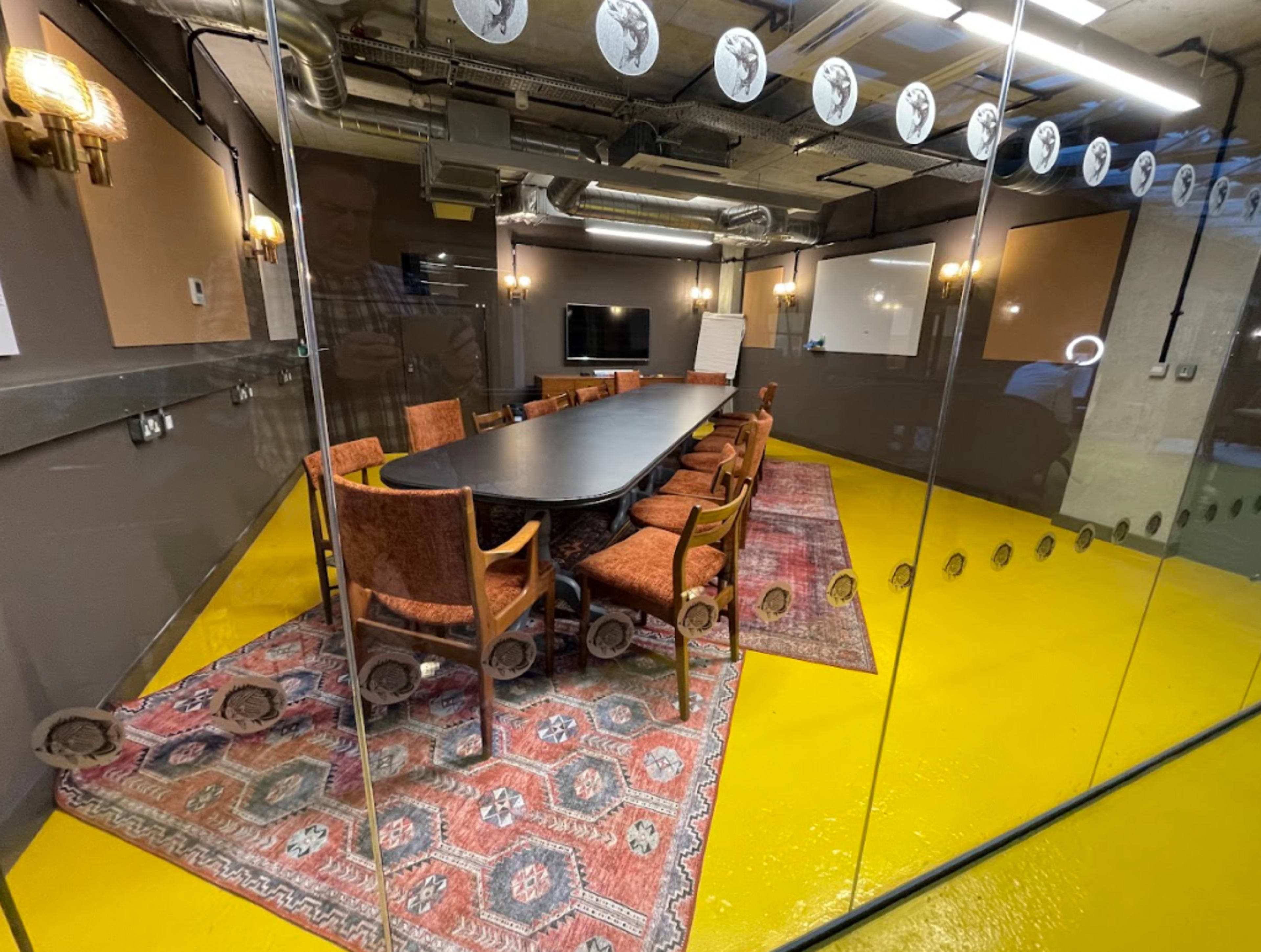A meeting room features a long table surrounded by upholstered chairs, with a large screen and whiteboards visible in a glass-walled space.