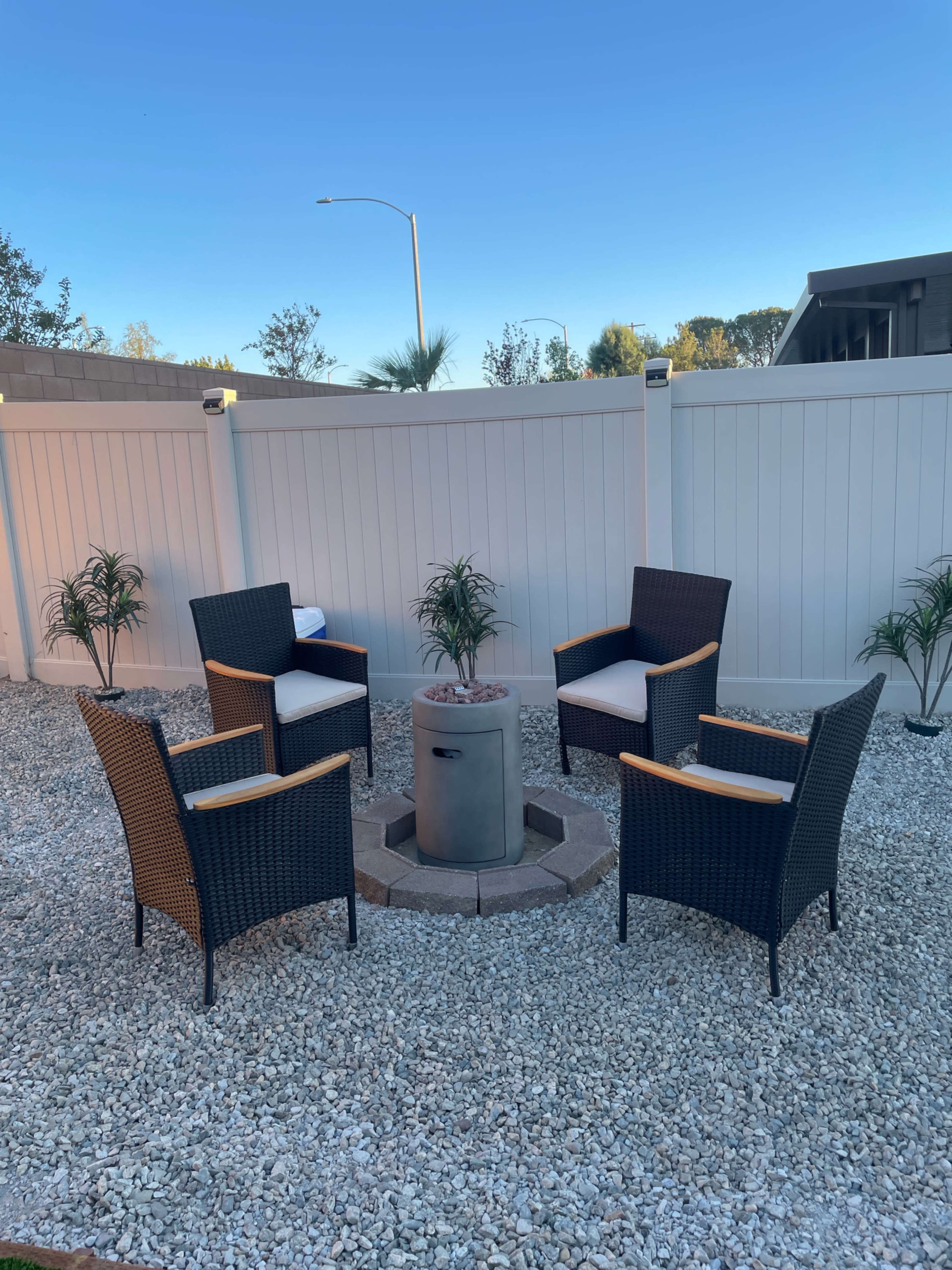 The image shows a circular seating arrangement with four wicker chairs around a stone fire pit, set in a landscaped area with gravel and a white privacy fence.