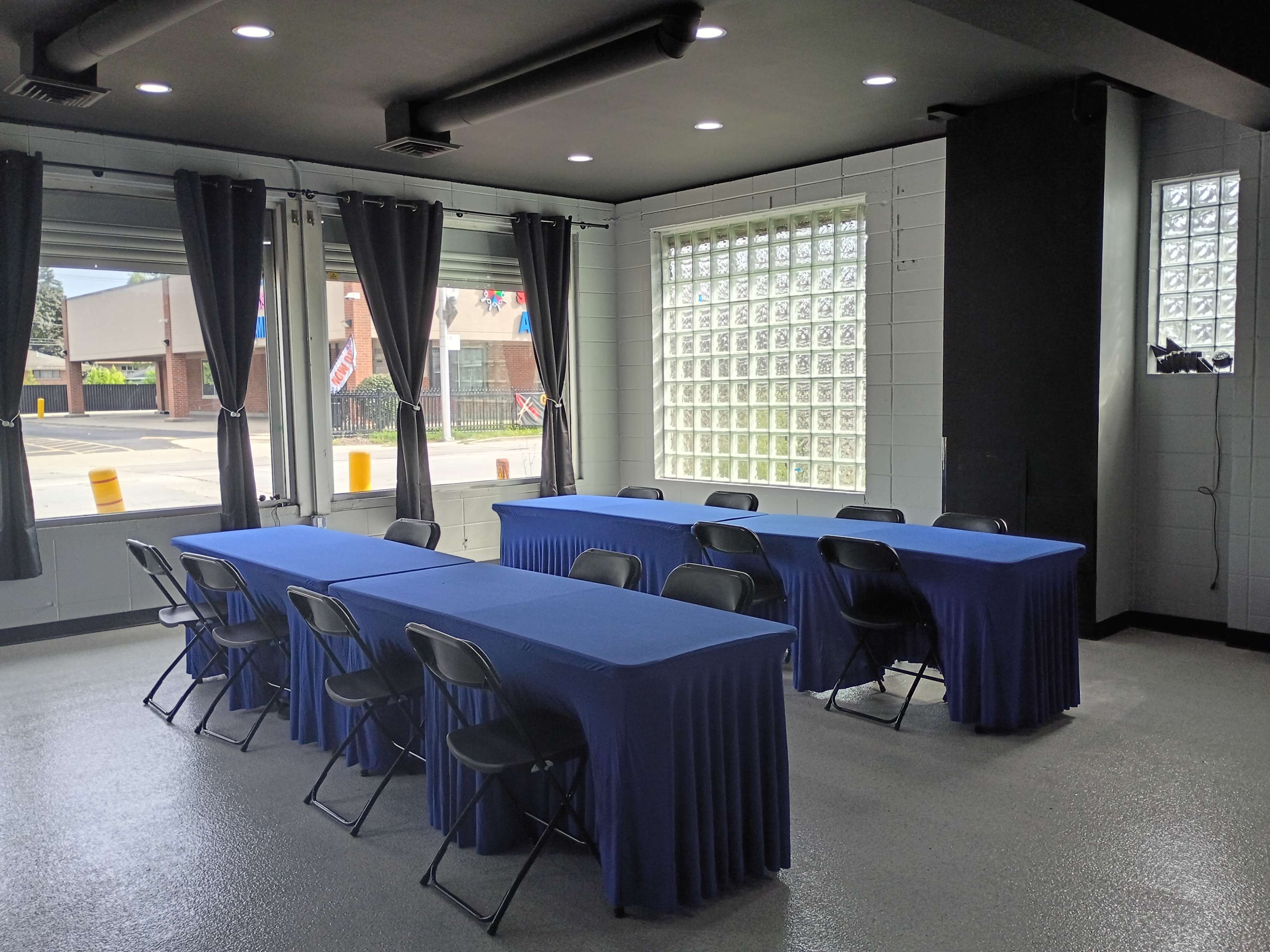 The image shows a meeting room with two rows of tables covered in blue cloth and black chairs arranged around them, with large windows allowing natural light to enter.
