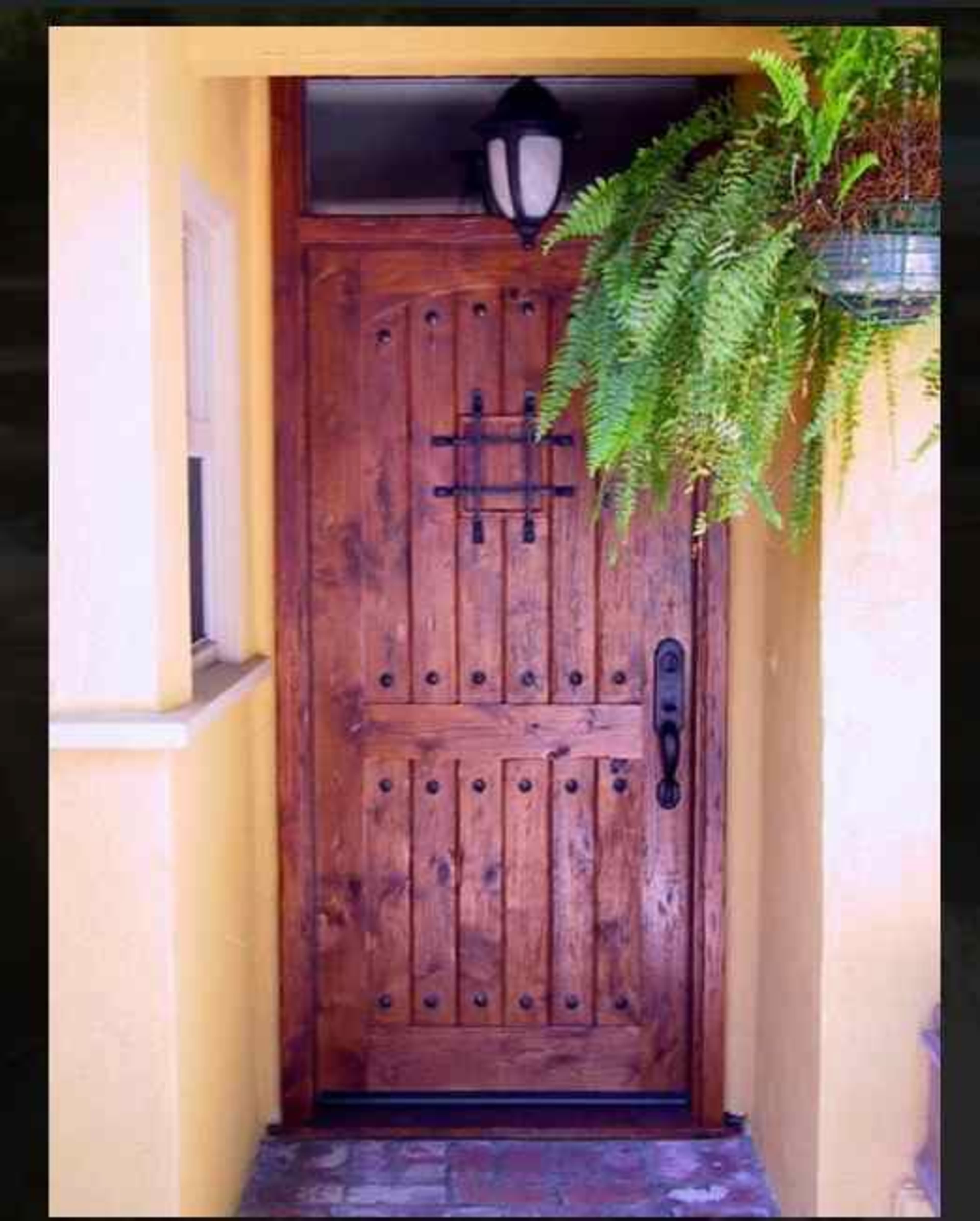 A wooden door with vertical planks and decorative metal accents is framed by a yellow wall and adorned with a hanging fern planter.