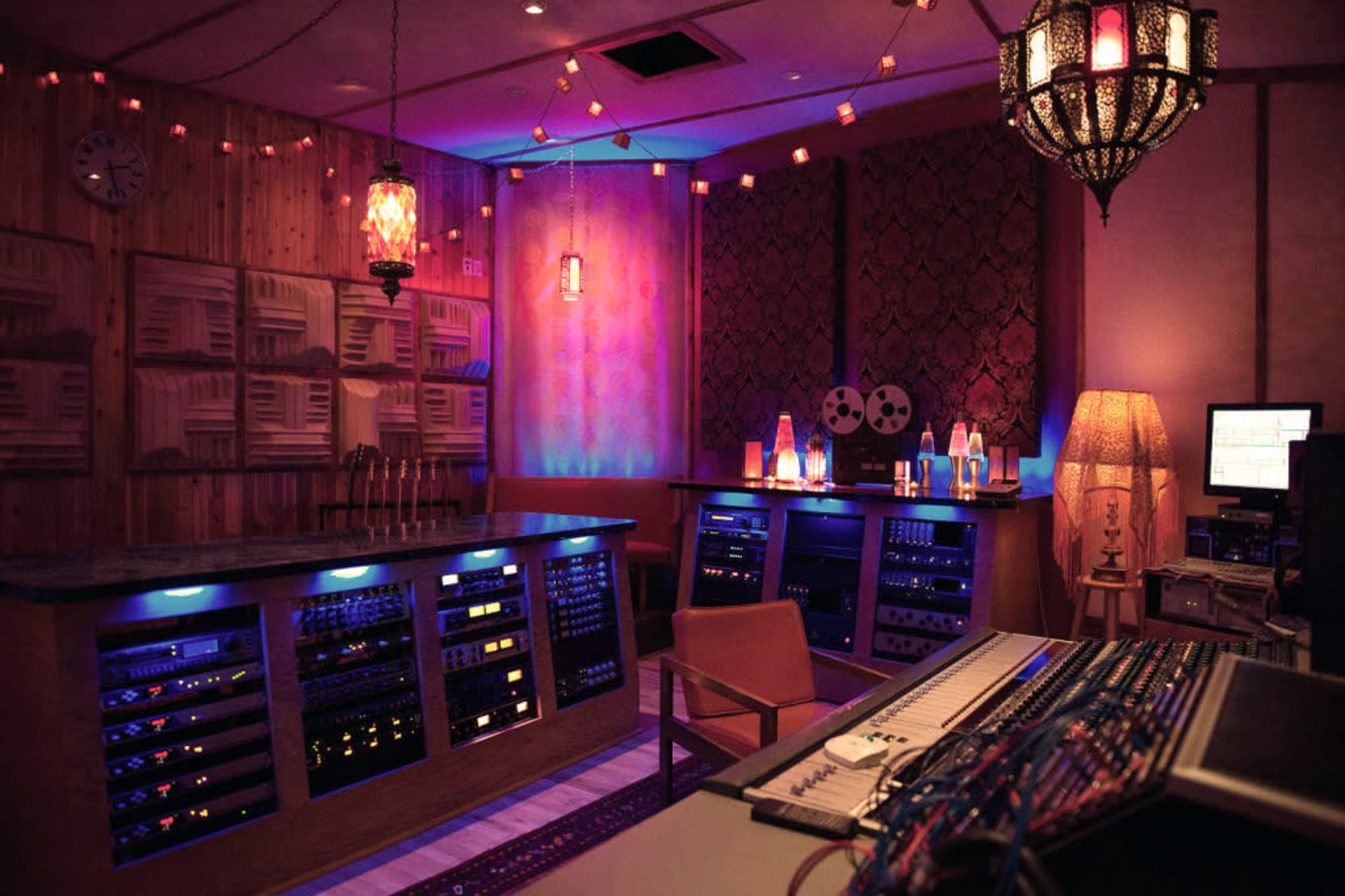 A dimly lit recording studio with various audio equipment, including a mixing console and tape reels, arranged alongside wooden paneling and decorative lighting.