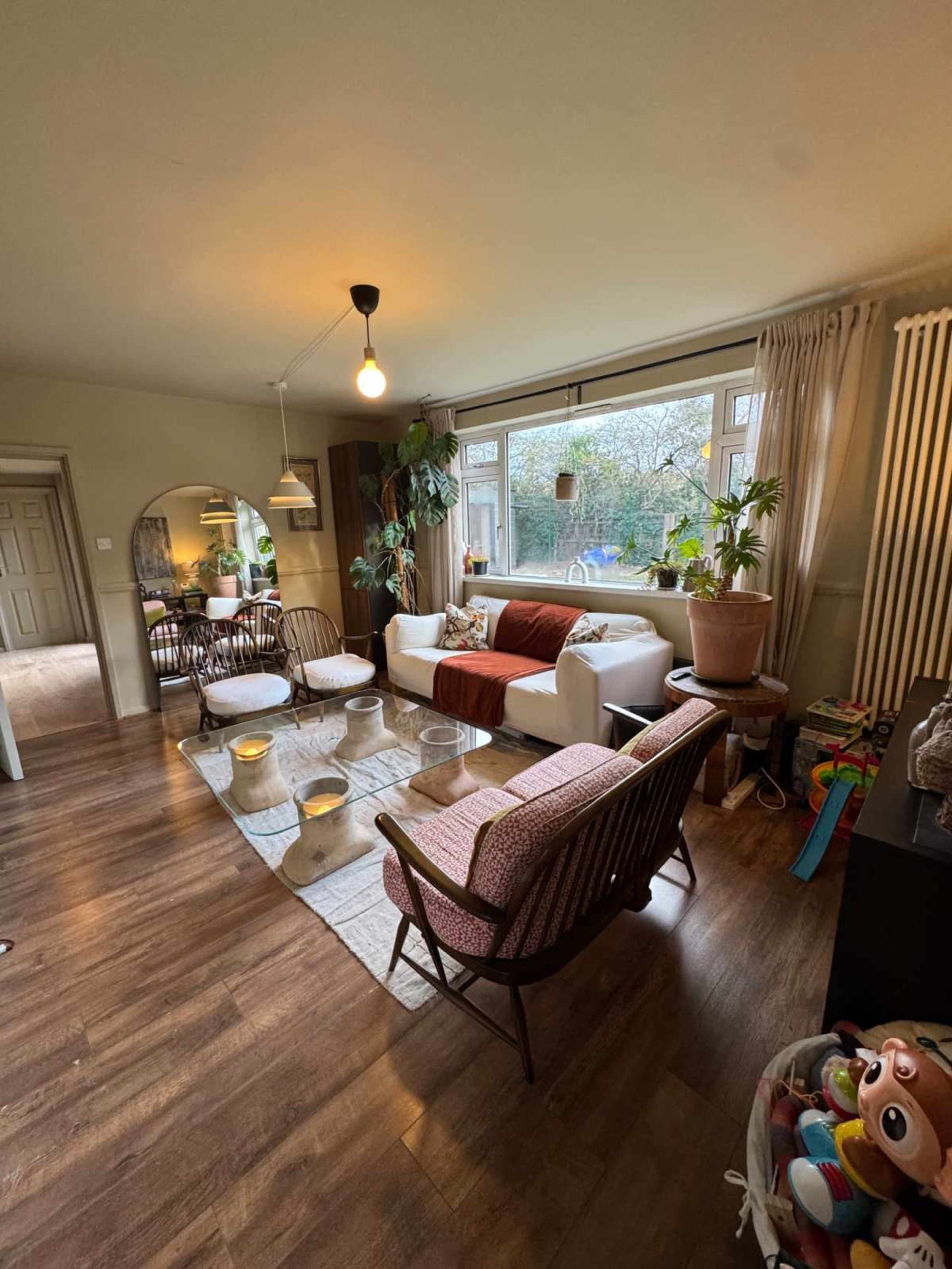 The image shows a cozy living room featuring a white sofa, a pink armchair, a glass coffee table, and large windows with plants.