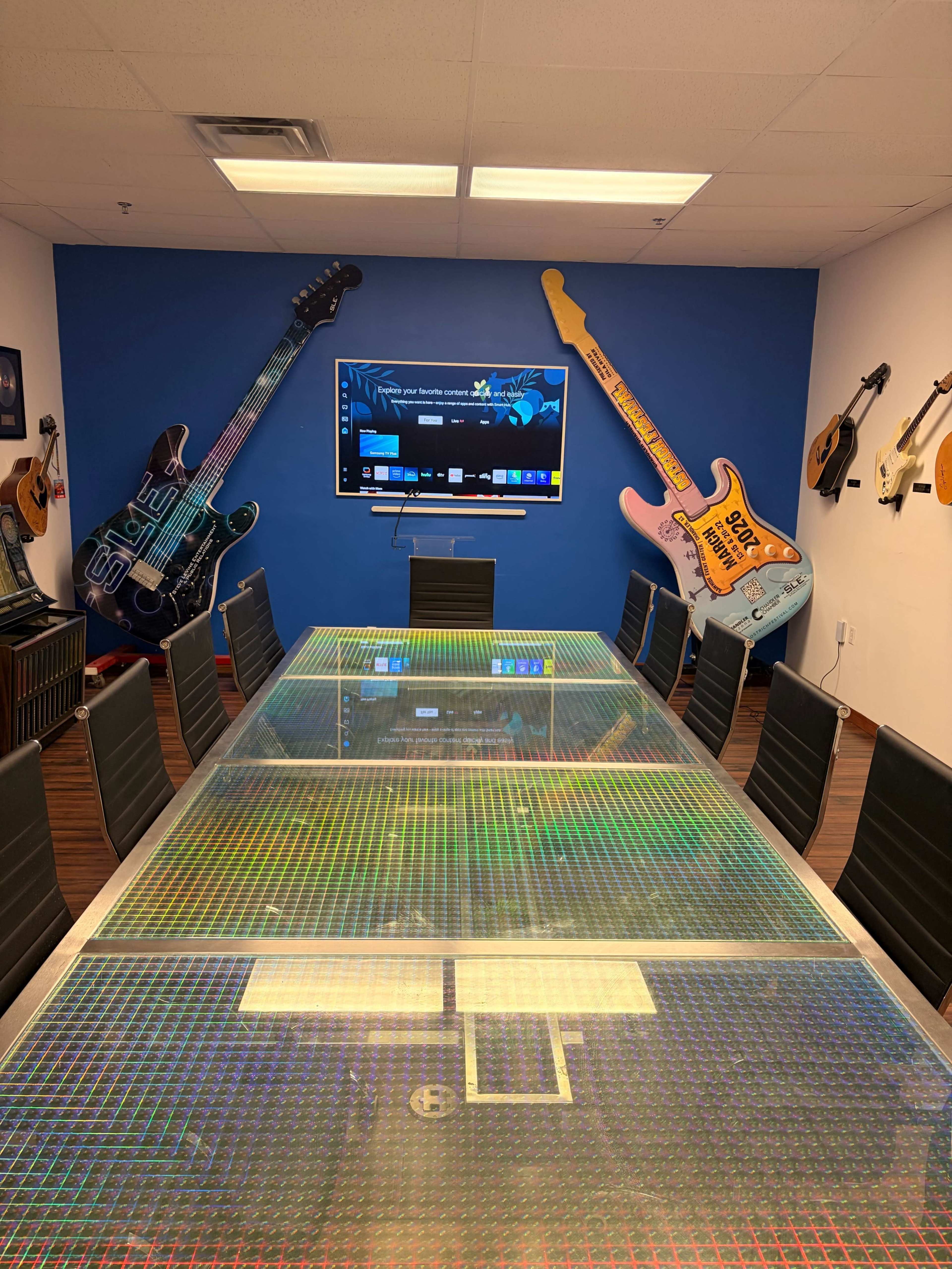 A modern conference room features a glass table surrounded by black chairs, with large electric guitar decorations on the walls and a television mounted in the center.