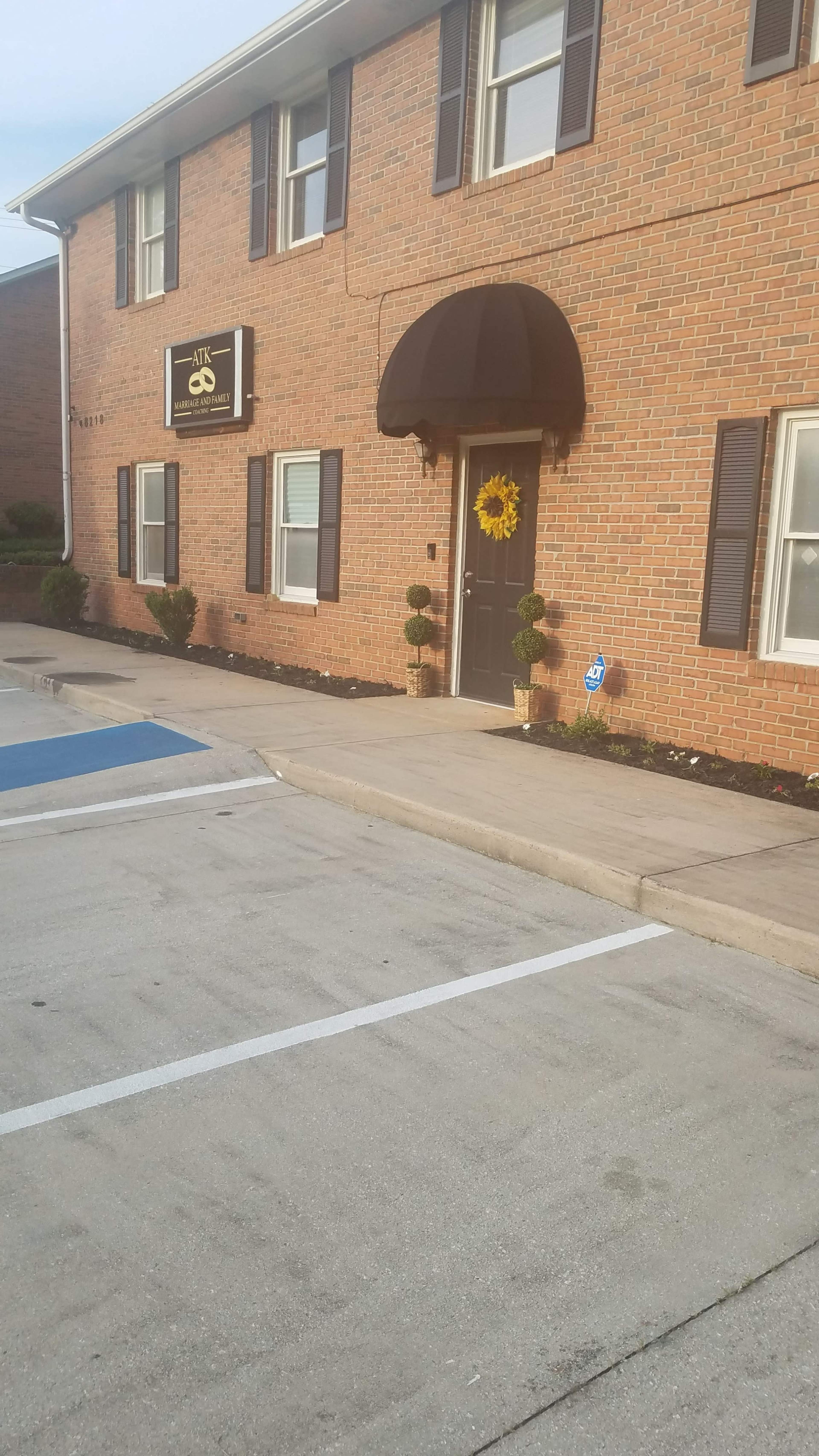A brick building with a dark awning above a door decorated with a sunflower, surrounded by neatly trimmed bushes and a parking area.