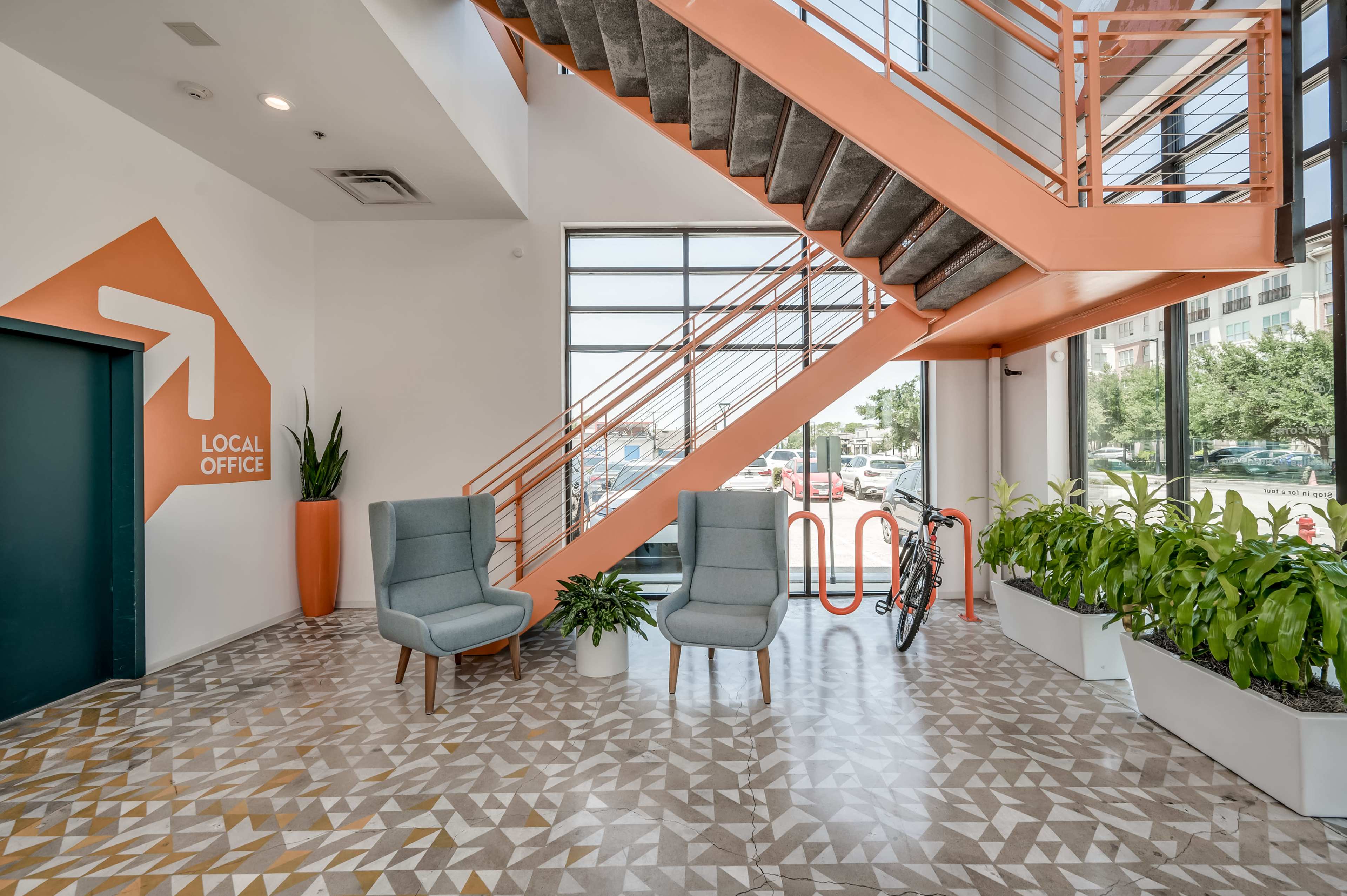 The image shows a modern entrance area featuring two grey chairs, a bicycle, and plants, with an orange staircase and a wall displaying a directional sign for a local office.