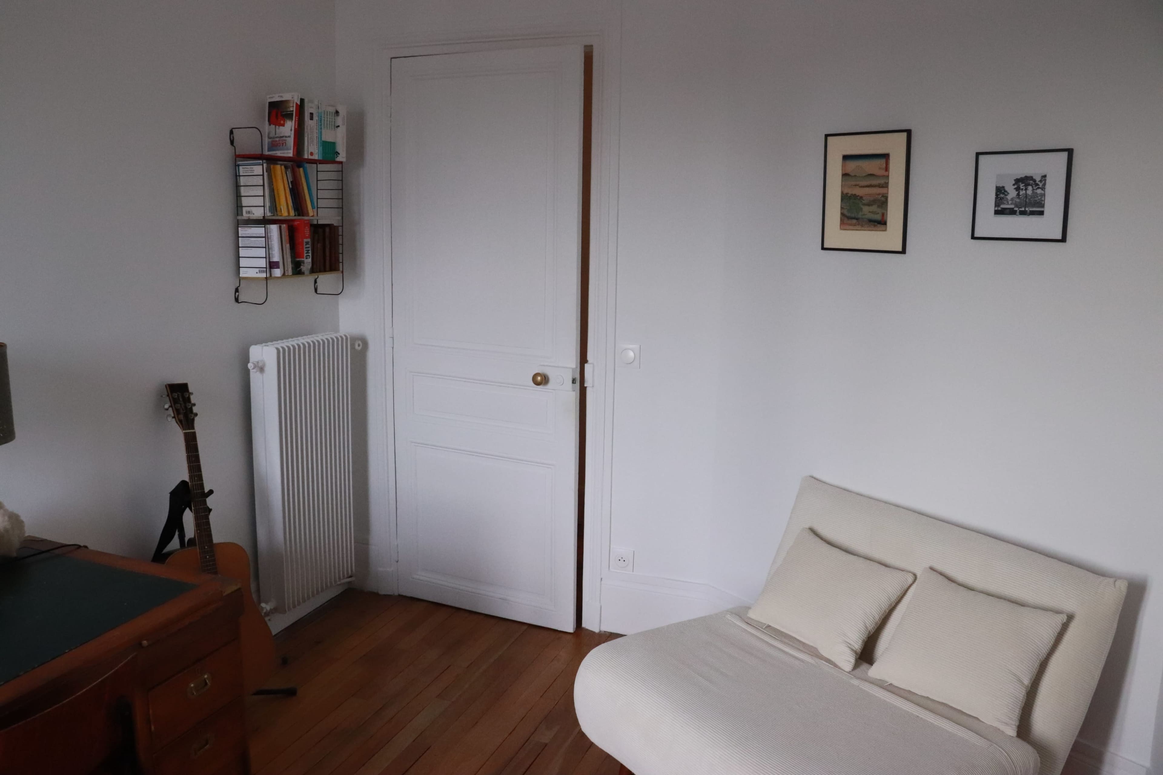 The image shows a minimalistic room featuring a white sofa, wooden flooring, a guitar leaning against the wall, a small desk, and framed pictures hanging on the wall.