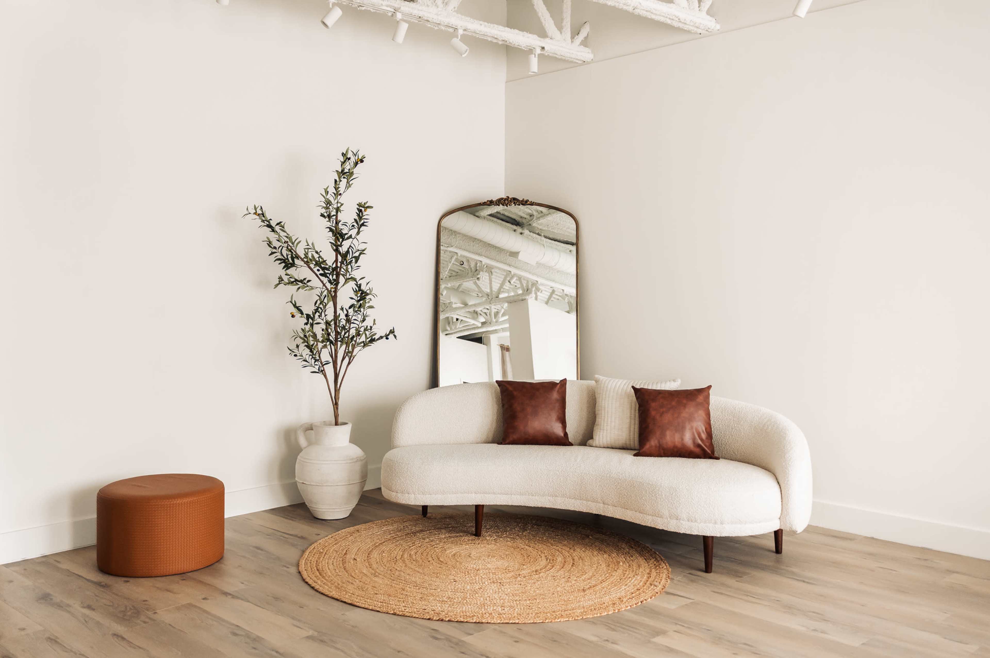 The image shows a modern, minimalist interior featuring a curved white sofa with two brown pillows, a round jute rug, a cylindrical orange ottoman, a potted tree, and a large mirror against a white wall.