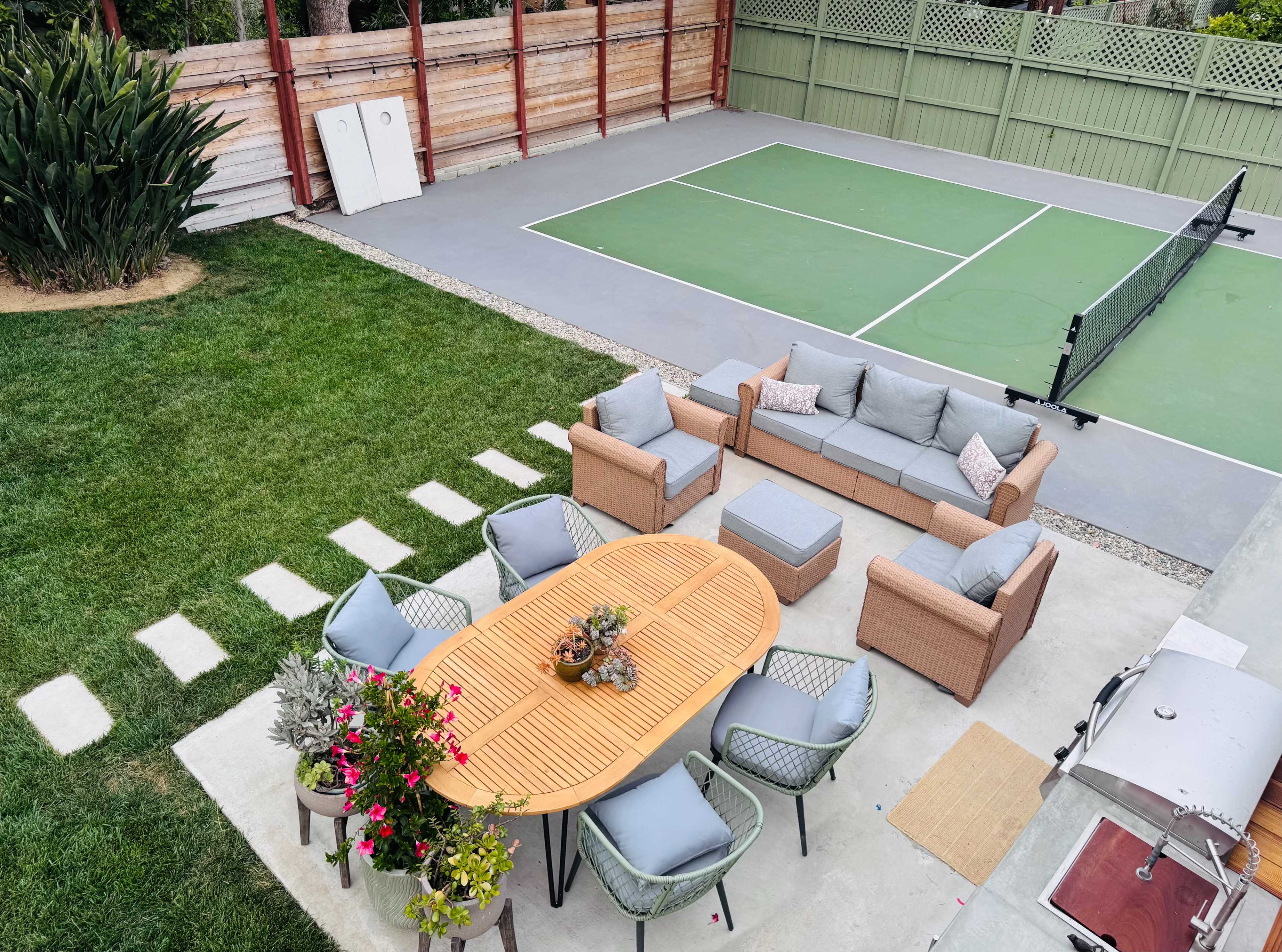 A backyard features a seating area with a round table and chairs, surrounded by green grass and a tennis court in the background.