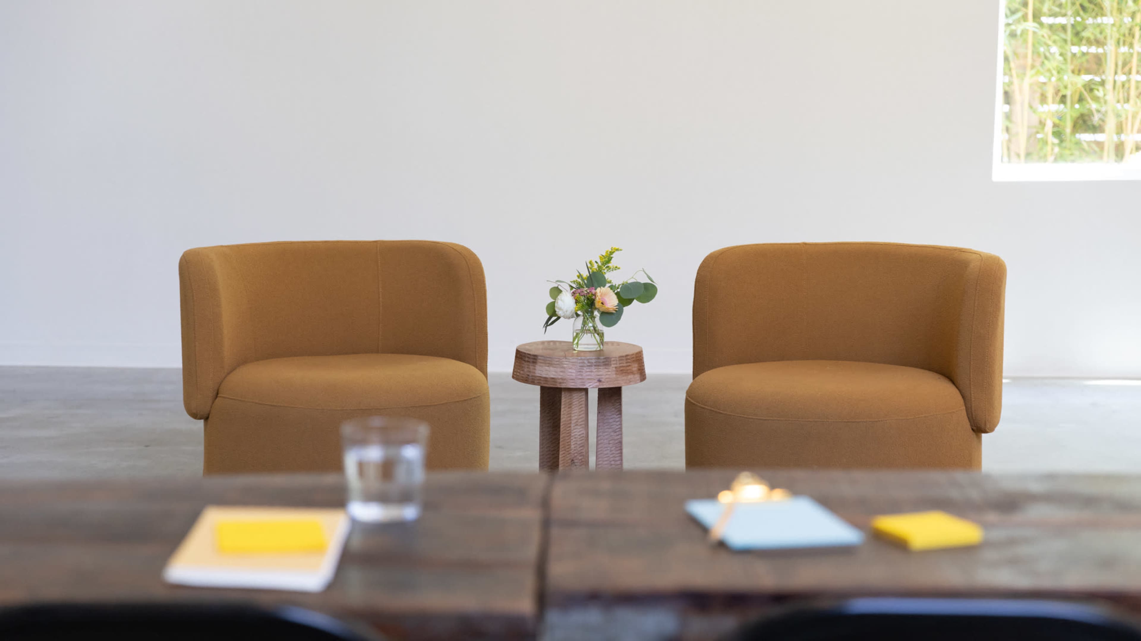 Two tan chairs are positioned facing each other near a small wooden side table with a vase of flowers, while a rustic table with notebooks and a glass of water is in the foreground.