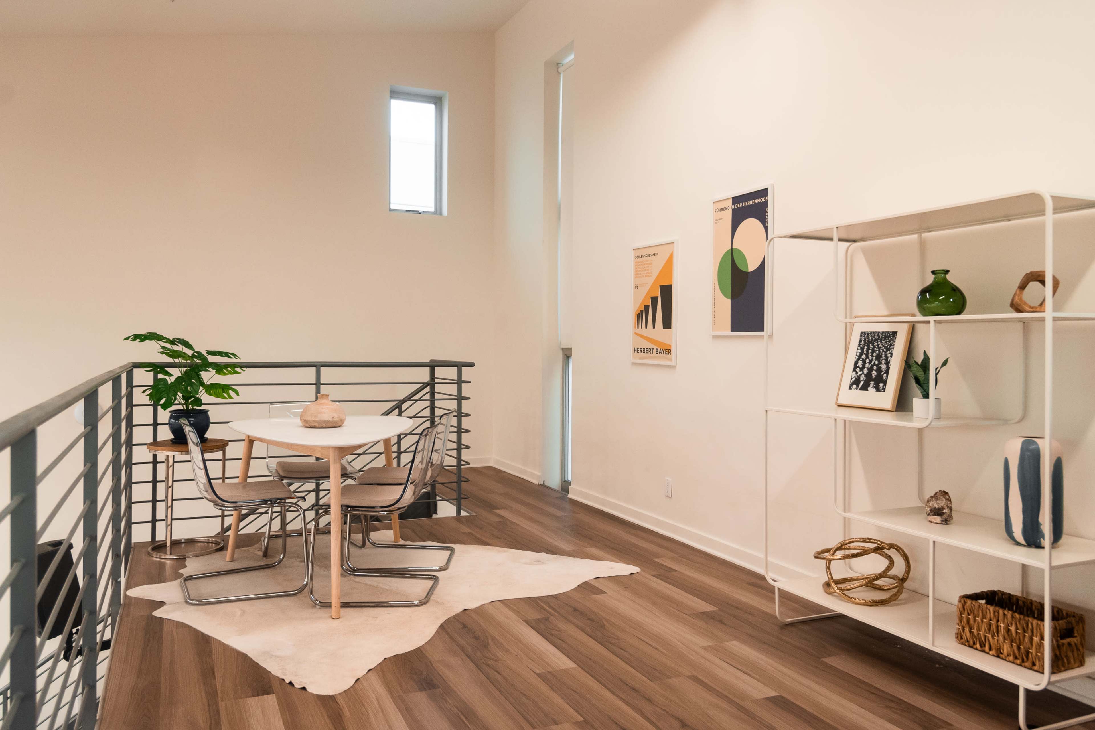 A modern, open-concept living space features a dining table surrounded by chairs, a decorative plant, and a bookshelf against a light-colored wall.