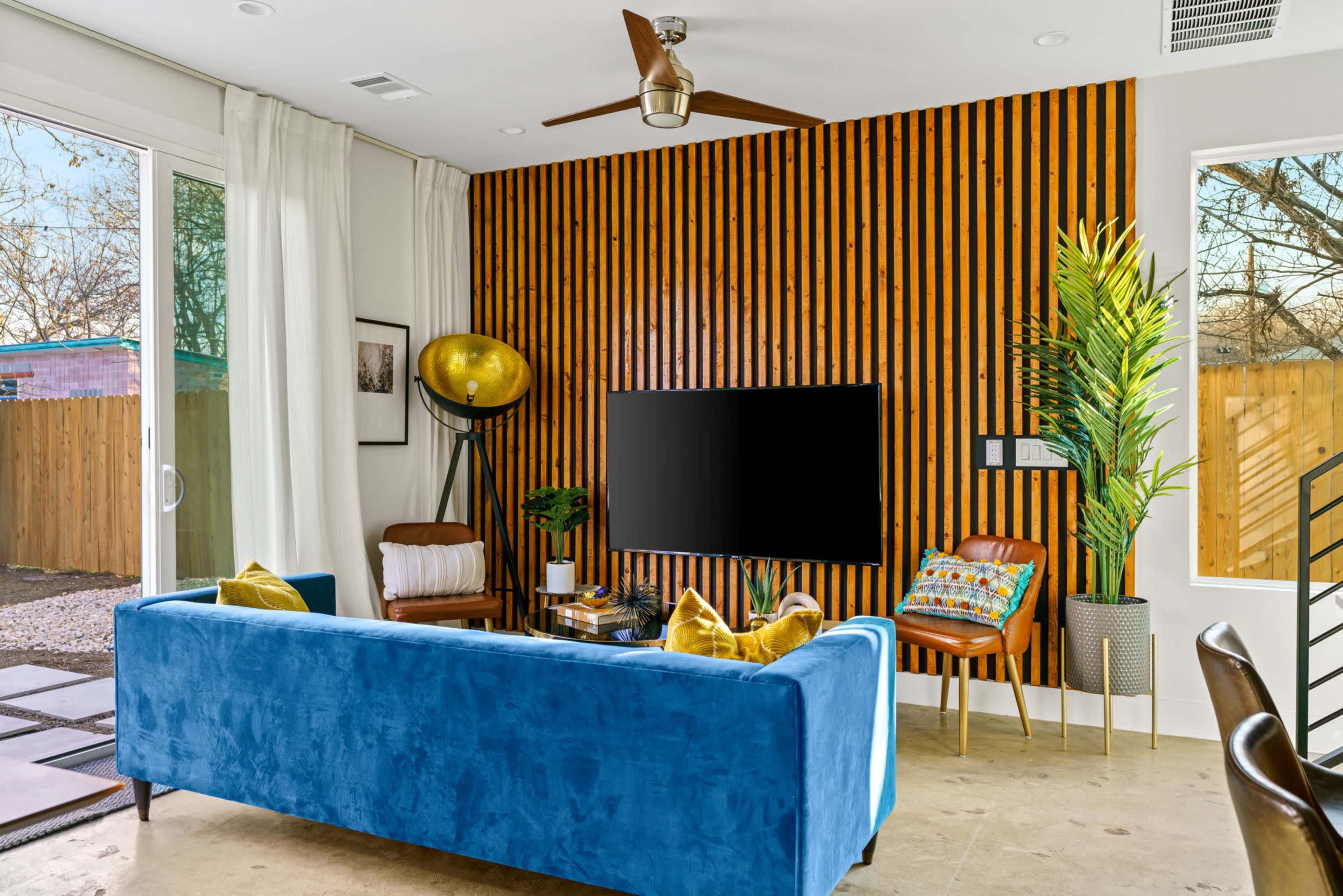 A modern living room features a blue sofa, wooden accent wall, and a television mounted above a low table, with large windows allowing natural light.