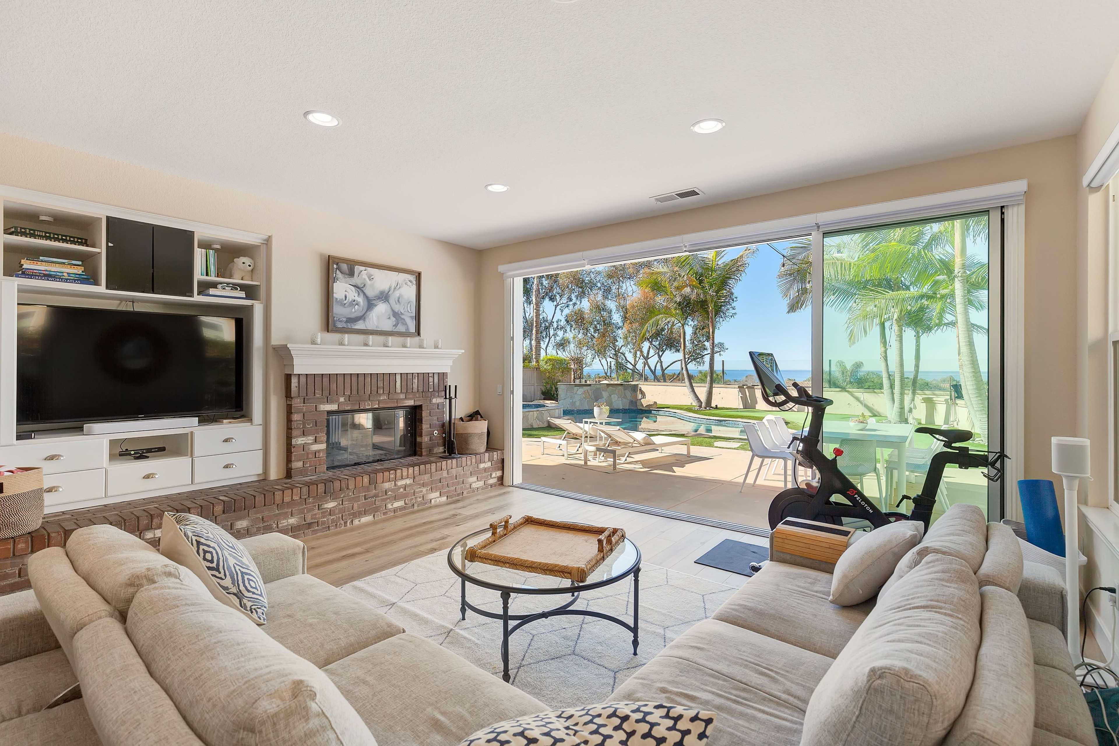 A spacious living room featuring a large window with a view of a patio and palm trees, along with a TV and a fireplace.