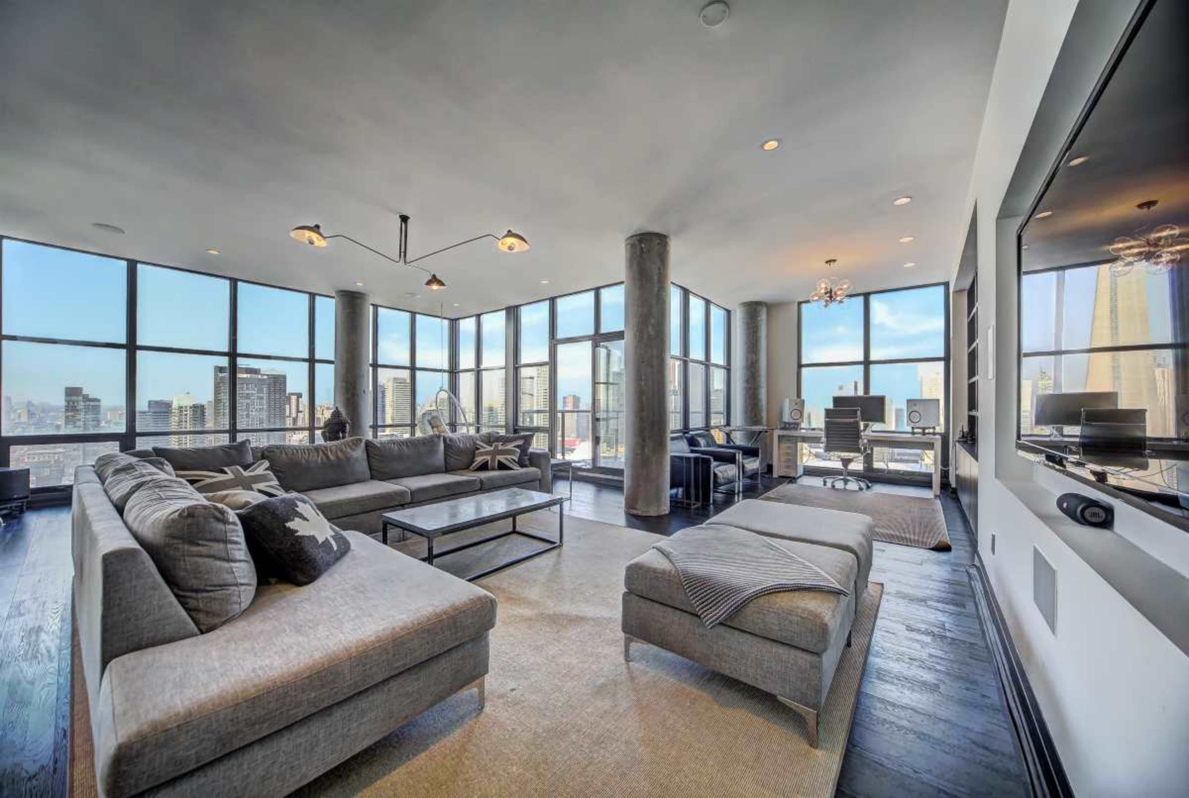 The image shows a spacious, modern living room with large windows that provide a view of a city skyline, featuring a sectional sofa, a coffee table, and a sleek entertainment area.