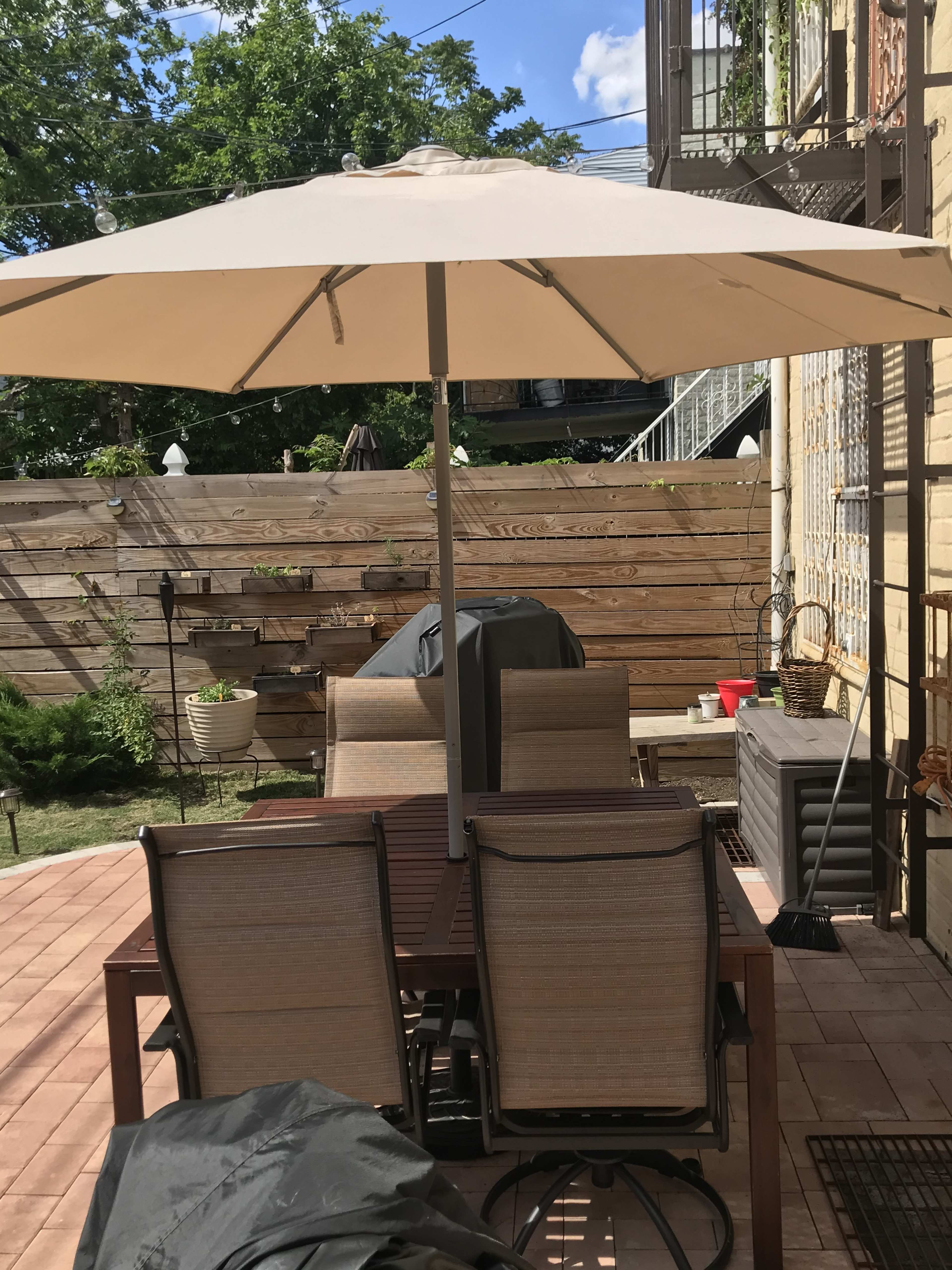 A patio area features a table with several chairs under an umbrella, surrounded by a wooden fence and potted plants.