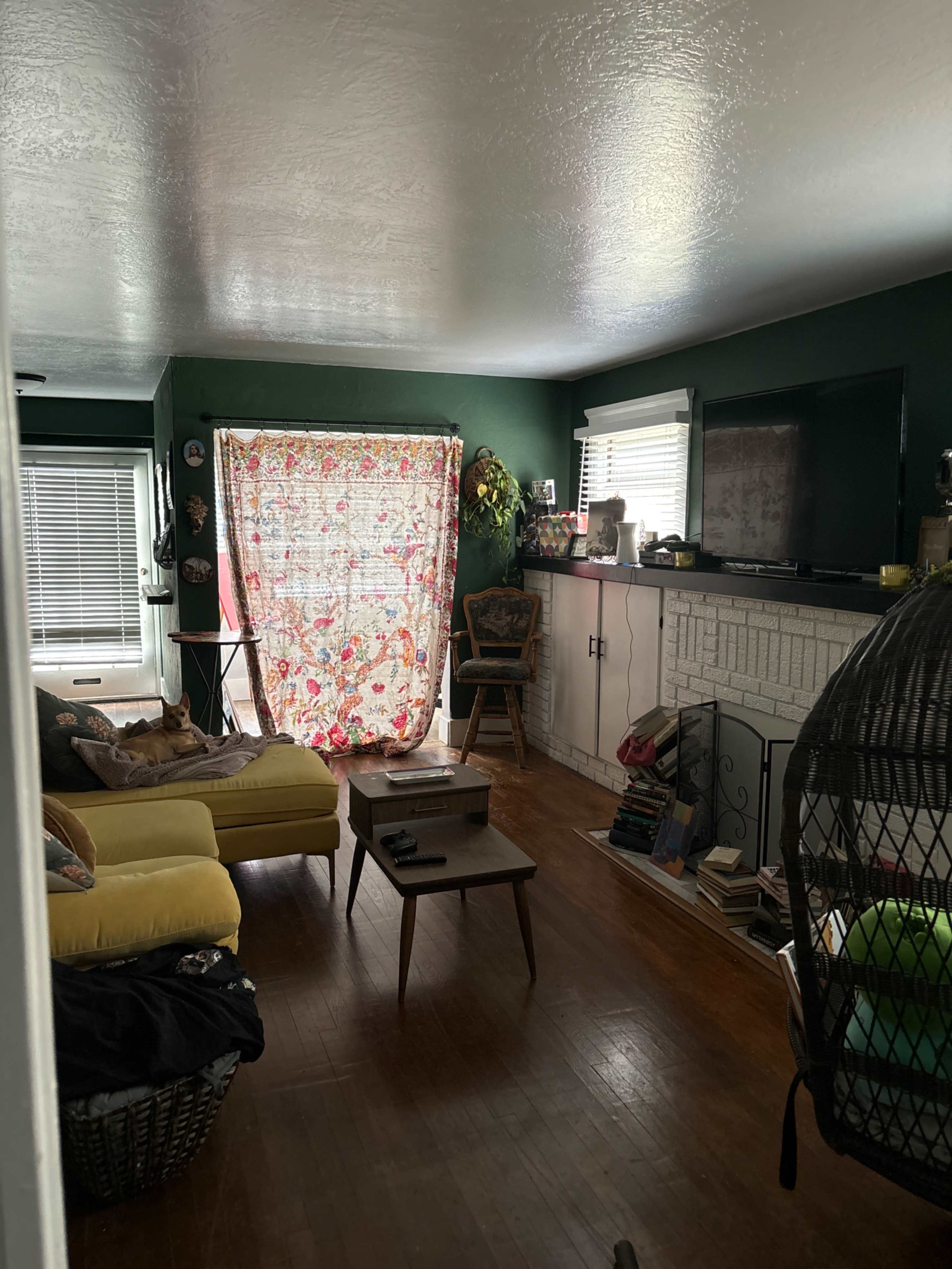 The living room features a yellow couch, a wooden coffee table, and a floral curtain covering a window, with bookshelves and a television situated along one wall.