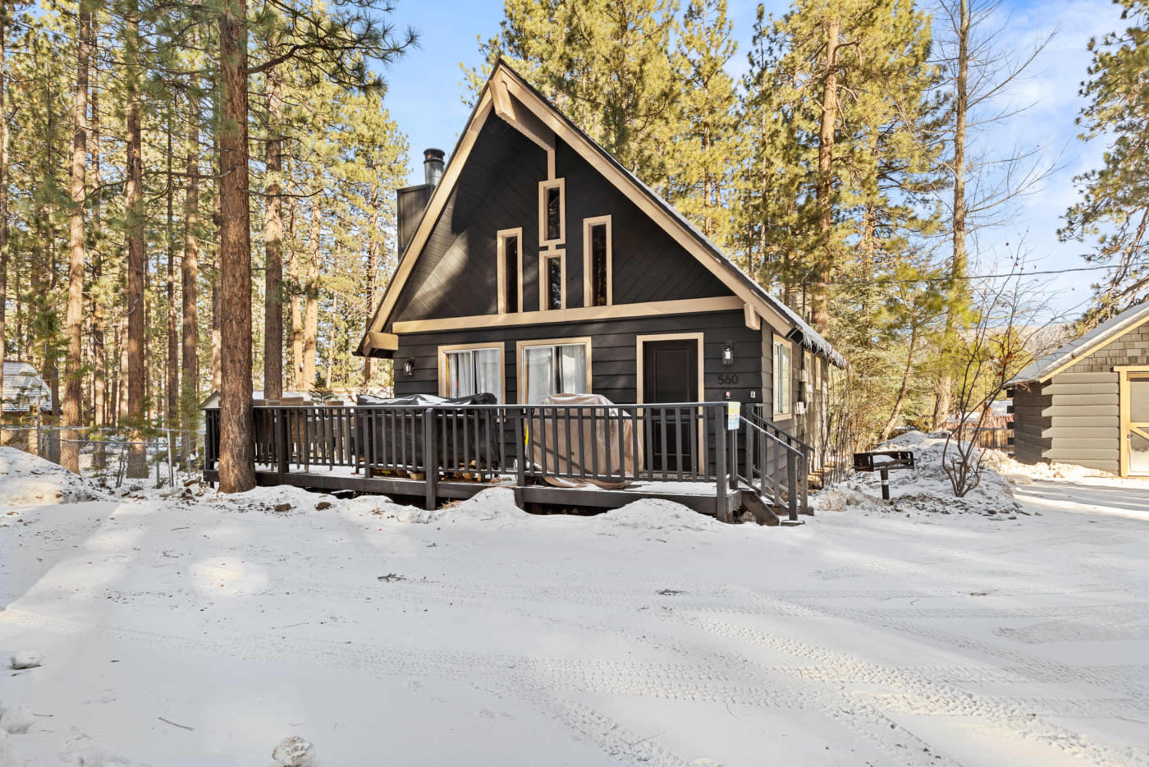 A cabin with a triangular roof and a front deck is situated among tall pine trees in a snowy landscape.