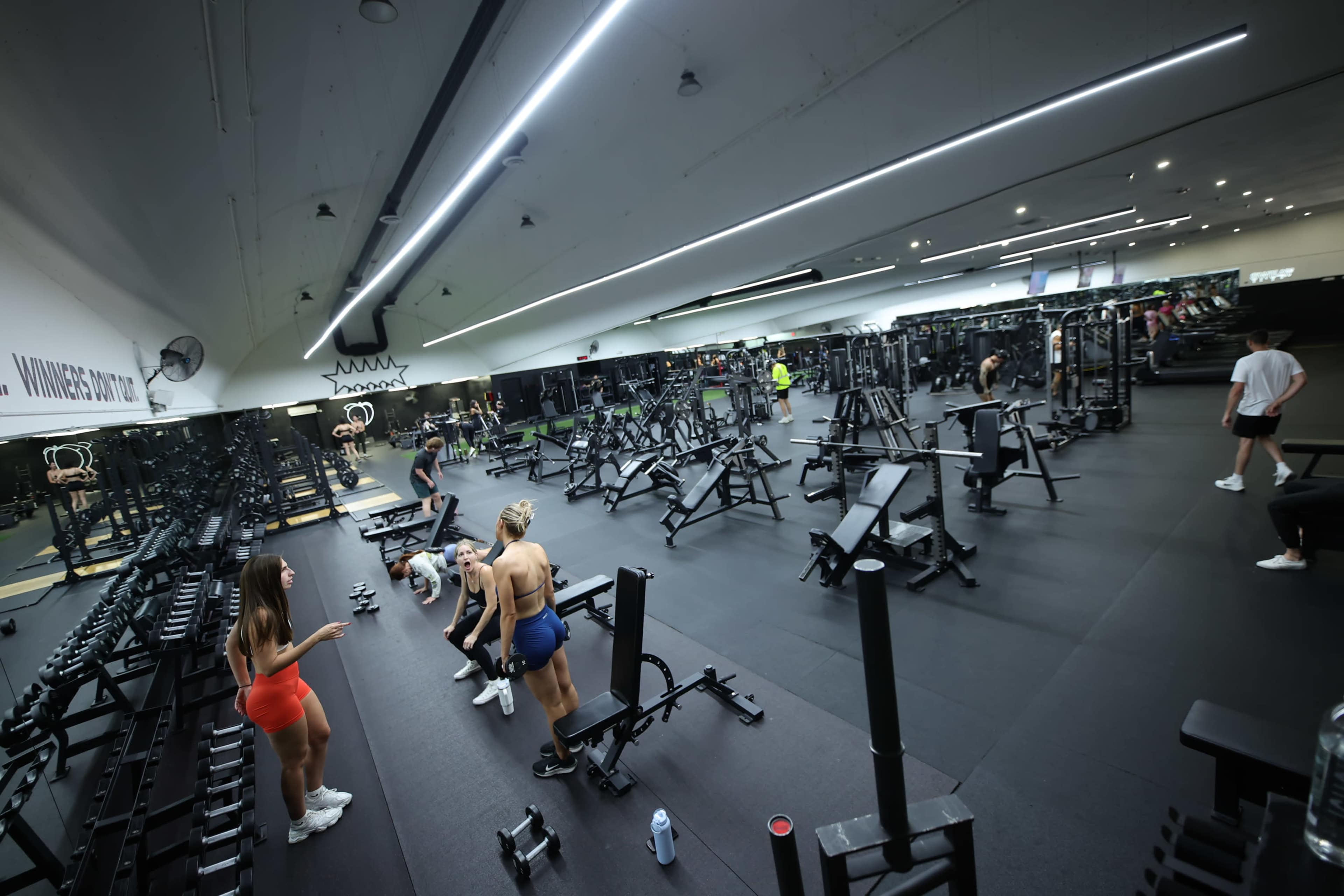 The image shows a spacious gym filled with various workout equipment, with several people engaged in exercise activities.