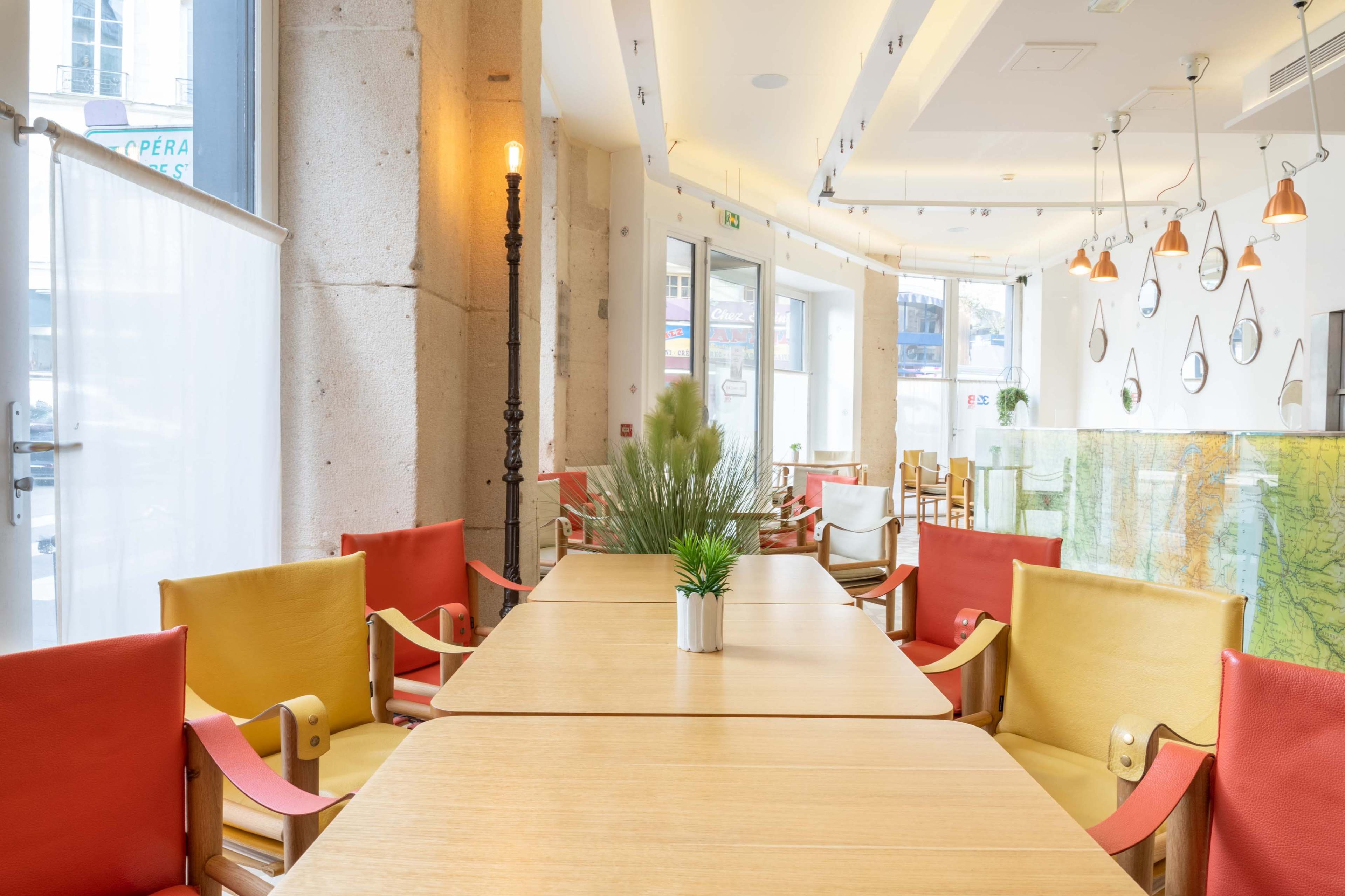 The image shows a modern restaurant interior with wooden tables, colorful chairs, and decorative plants.