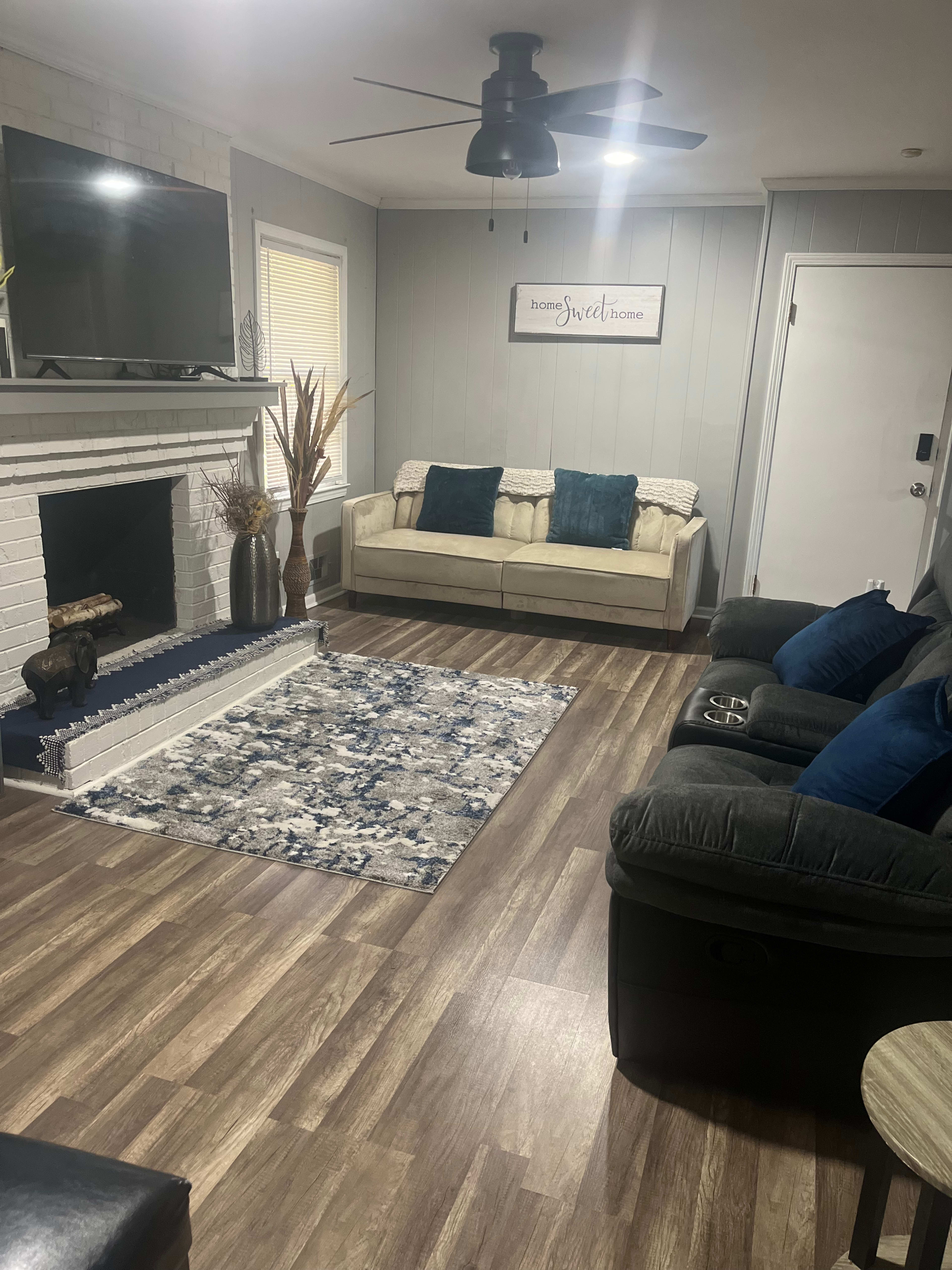 The living room features a light-colored sofa with blue cushions, a dark recliner, a patterned rug, and a television mounted above a white brick fireplace.
