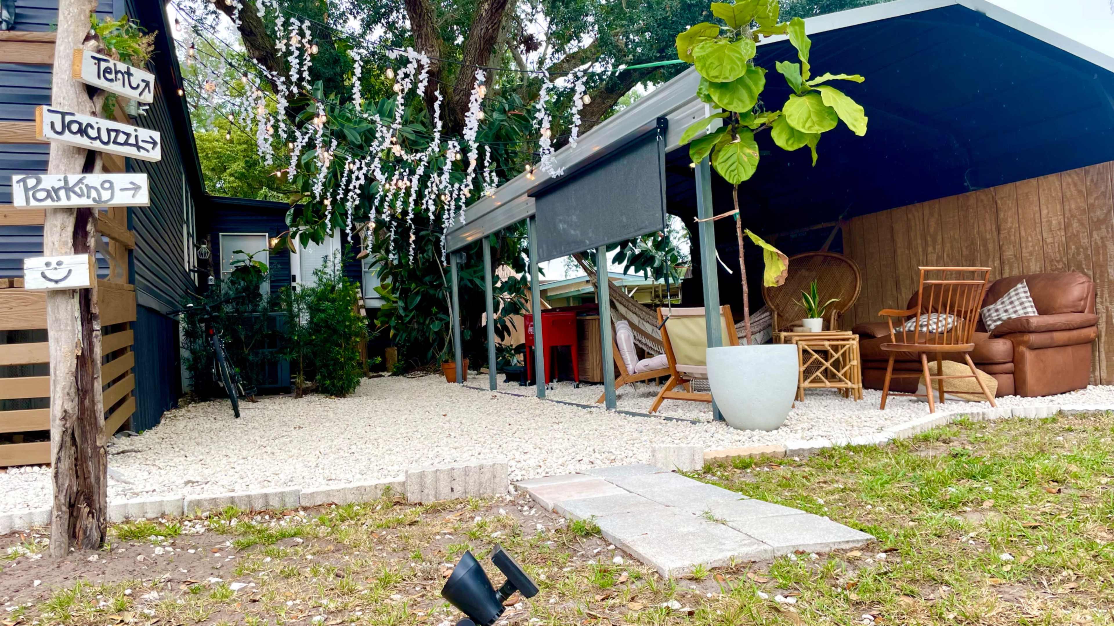 A landscaped outdoor space featuring a covered seating area with furniture, decorative plants, and gravel flooring, alongside a sign indicating tent and jacuzzi locations.
